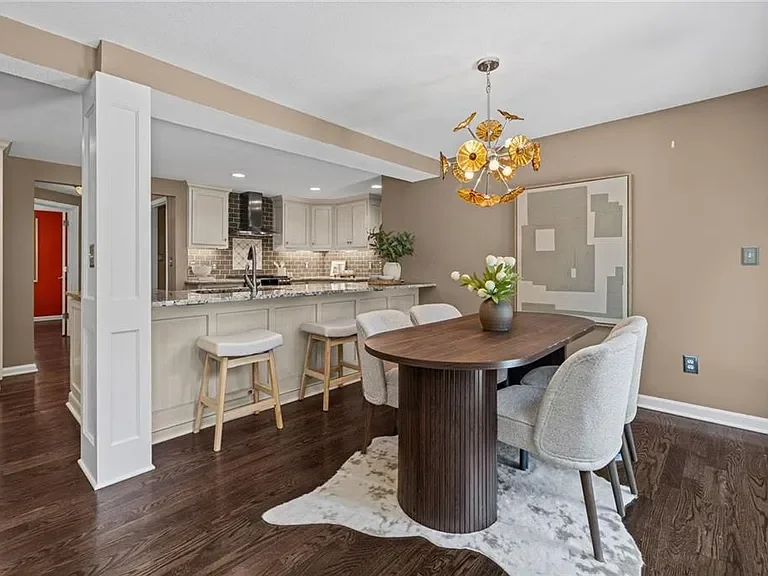 Open-concept dining area with a dark wood oval table, six upholstered chairs, and a modern chandelier. Adjacent kitchen with white cabinets, subway tile backsplash, and bar stools.