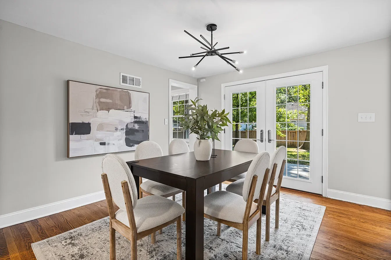 Dining room with a dark wood table, six cream-colored cushioned chairs, a white vase with green leaves, a modern black chandelier, and glass double doors leading outside, with a gray area rug and light gray walls.