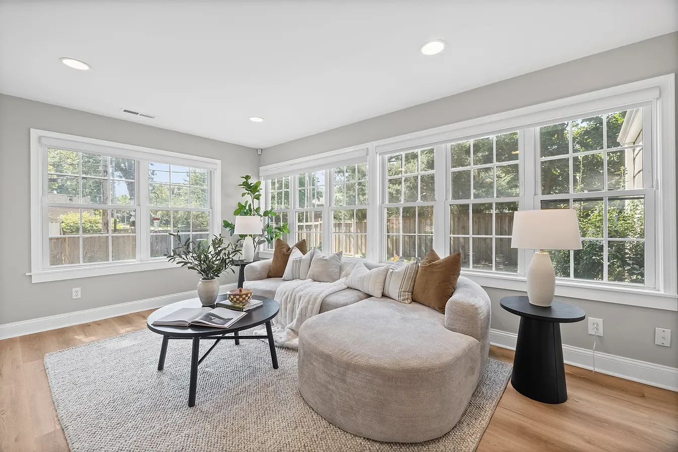 Bright living room with large windows, beige sectional sofa with throw pillows, black coffee table with open book and bowl, black side table with white lamp, potted plants, light wood flooring, and a neutral color palette.