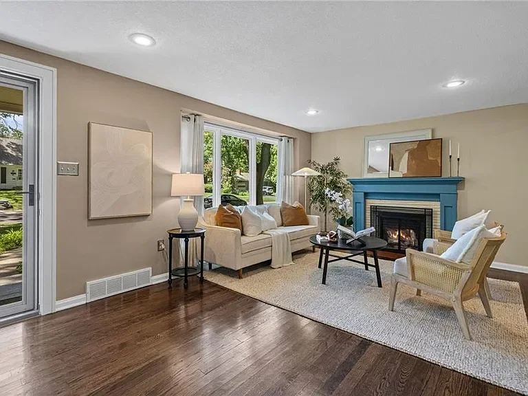 A well-lit living room with a beige wall, hardwood floor, beige couch with cushions, armchair, coffee table, fireplace with a blue mantel, and a window with view of a green lawn outside.