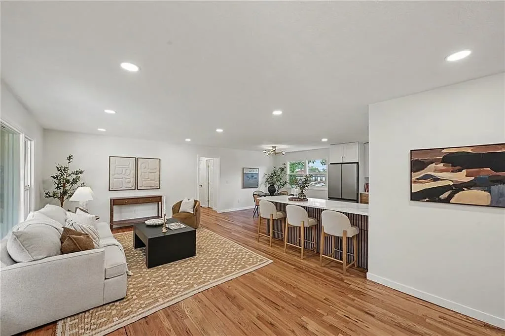 Open-concept living room and kitchen with white walls, hardwood floors, white sofa, armchair, rugs, artwork, and bar seating.