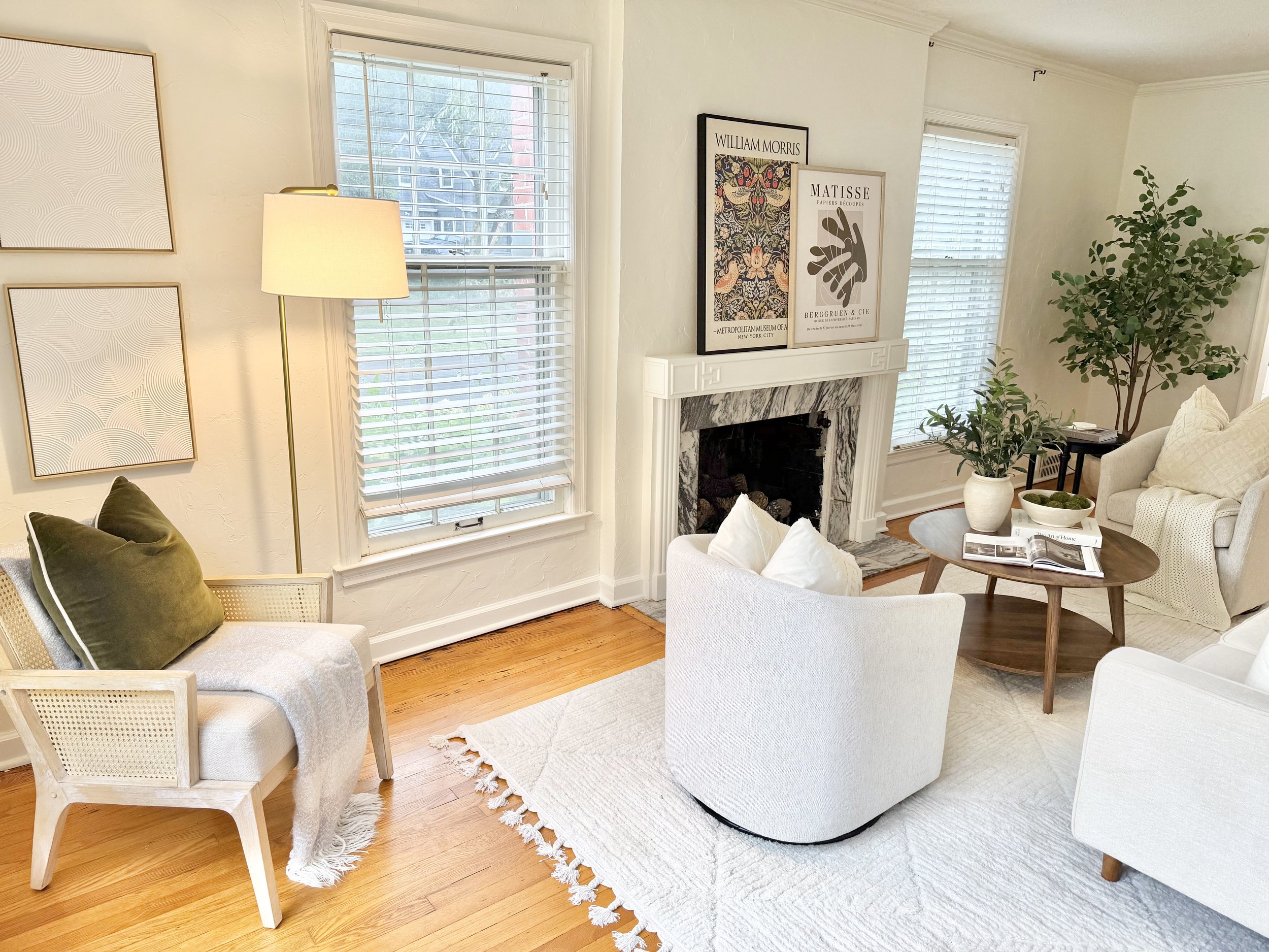 A cozy living room with a window, framed artwork, a white fireplace, a potted plant, a cream-colored couch, a marble-topped coffee table with magazines and bowls, and a white armchair with pillows.