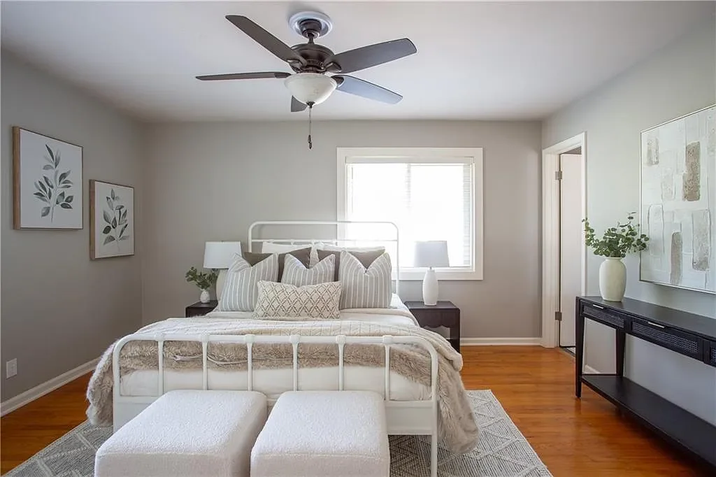 A bedroom with a white metal bed frame, neutral-colored bedding, two bedside tables with lamps, and a window behind the bed. There are wall art, a ceiling fan, a black console table with a vase and plant, and two white ottomans at the foot of the bed