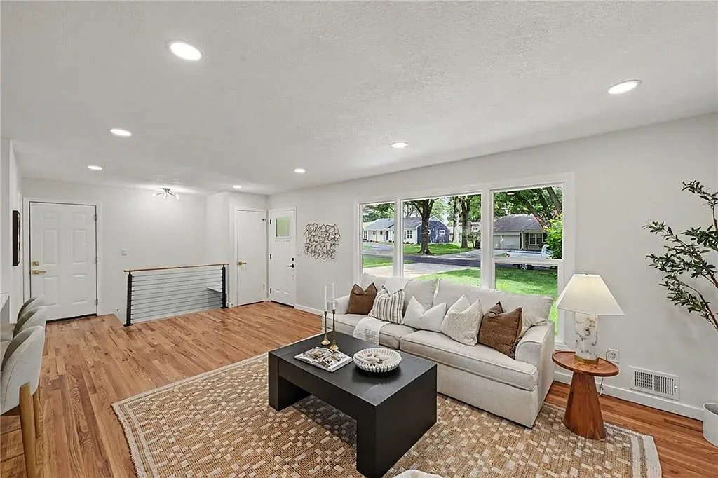 Living room with hardwood floors, white walls, large window, cream-colored sofa with pillows, side table with lamp, black coffee table, area rug, and chairs to the left.