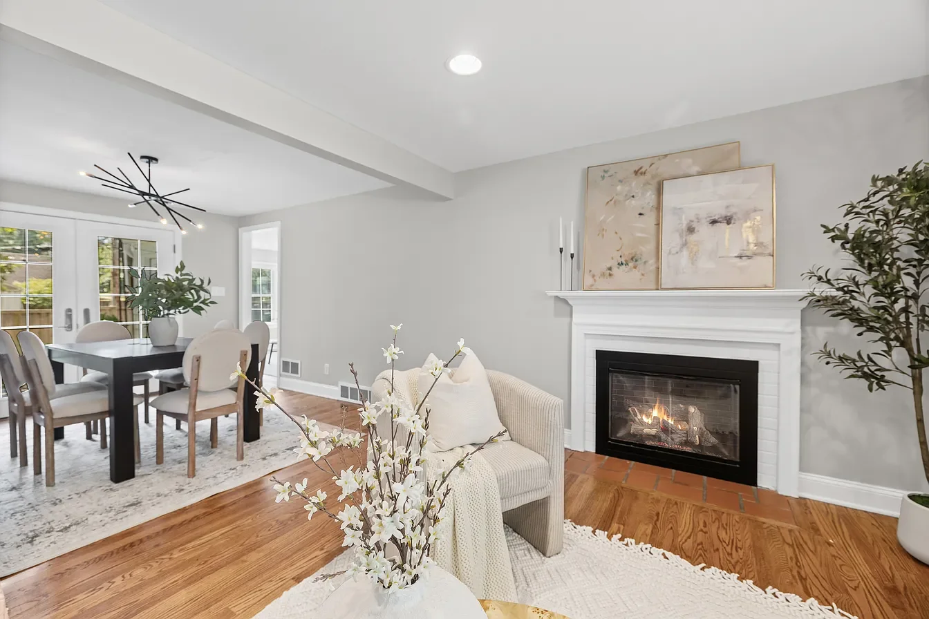 Living room with white walls, hardwood floors, a fireplace with white mantel and artwork above, a beige armchair with pillows, a glass vases with white flowers, and a dining area with a dark table and white chairs.