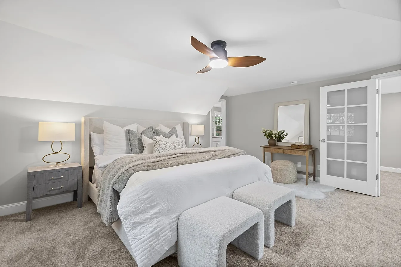A modern bedroom featuring a bed with white and gray bedding, two bedside lamps, a wooden ceiling fan, a bedside table, a mirror, a vase with flowers, and a pair of white ottomans in front of the bed, with double glass-paneled doors leading to anothe