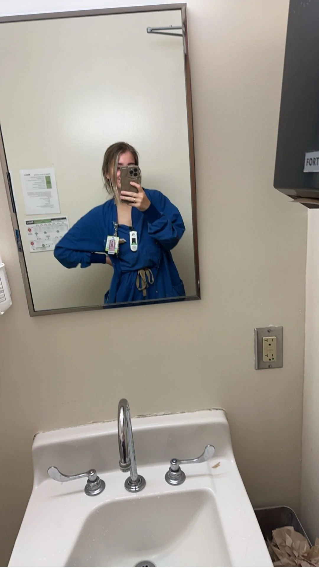 A person Labor and delivery Nurse wearing a blue medical gown taking a selfie in a hospital or clinic bathroom mirror, with a sink and wall-mounted outlets visible.