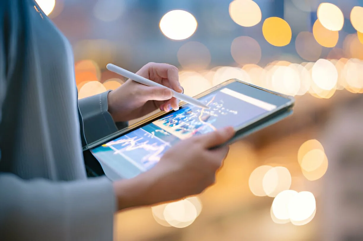 Person holding a tablet with a stylus in front of blurred city lights.