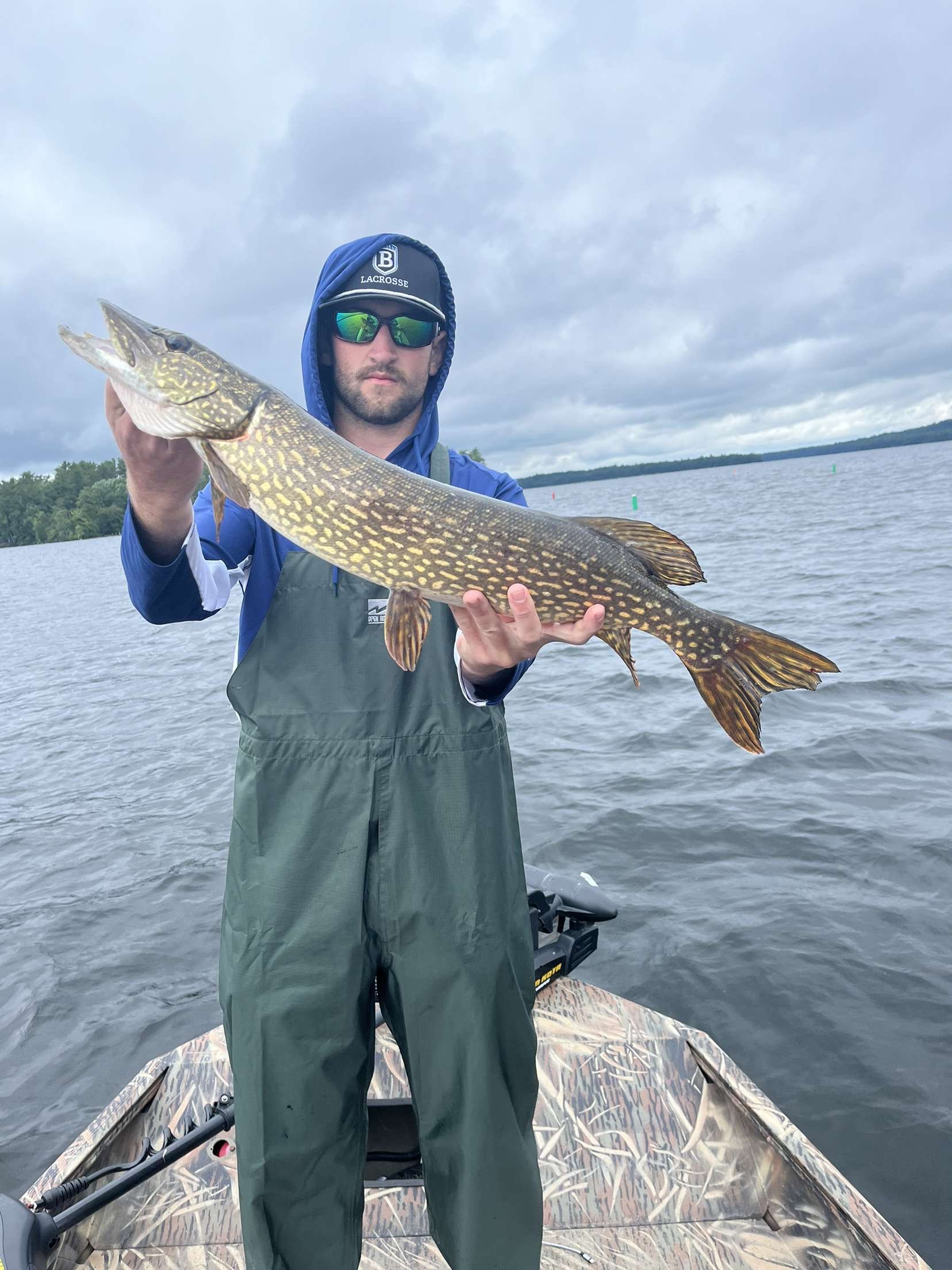 Man holding a large fish on a boat with a lake and cloudy sky in the background.