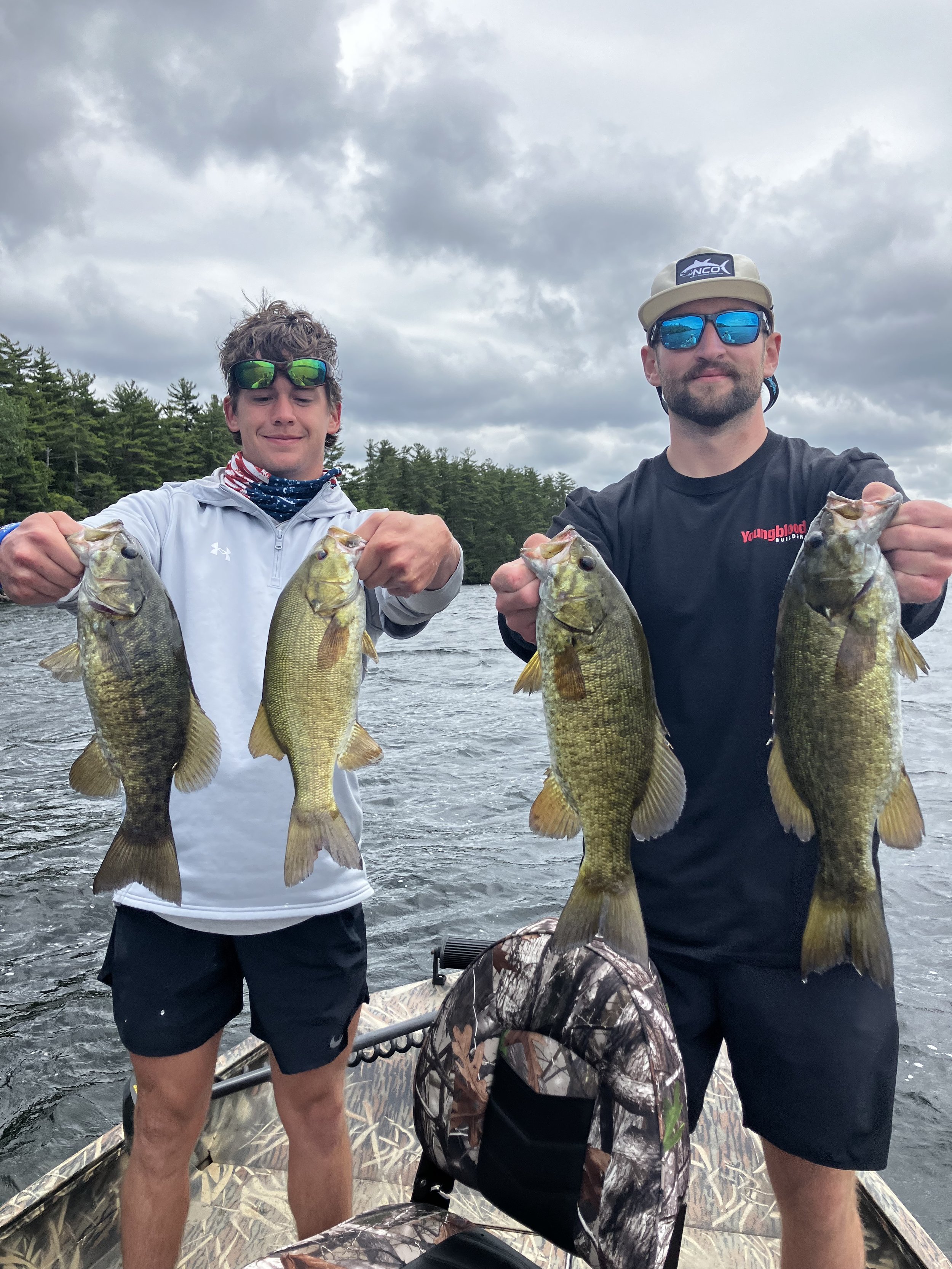 Two young men holding four fish each on a boat with a body of water and a forested shoreline in the background on a cloudy day.