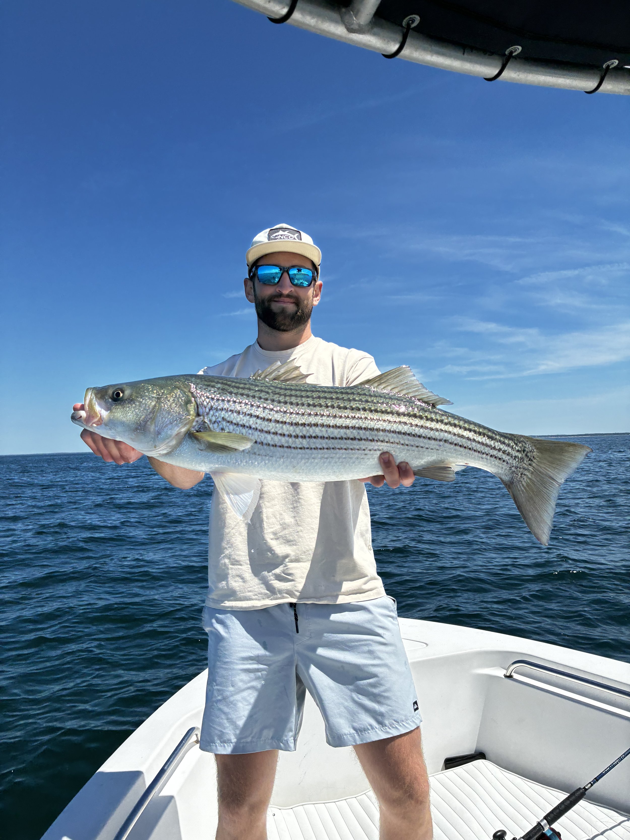 Man holding a large striped bass fish on a boat in the open ocean under a clear blue sky.
