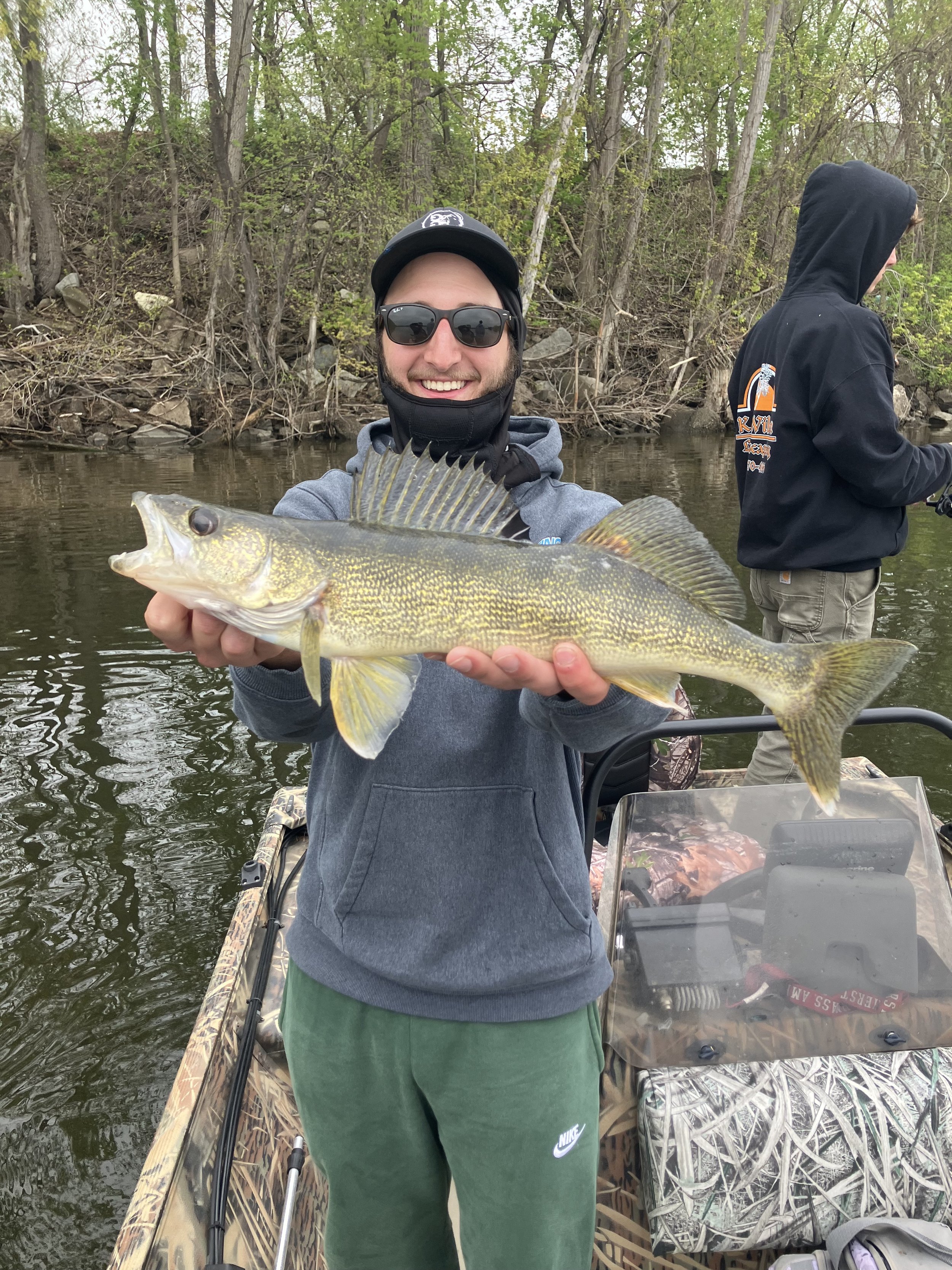 A man wearing sunglasses and a hoodie holding a large fish on a boat in a wooded river.
