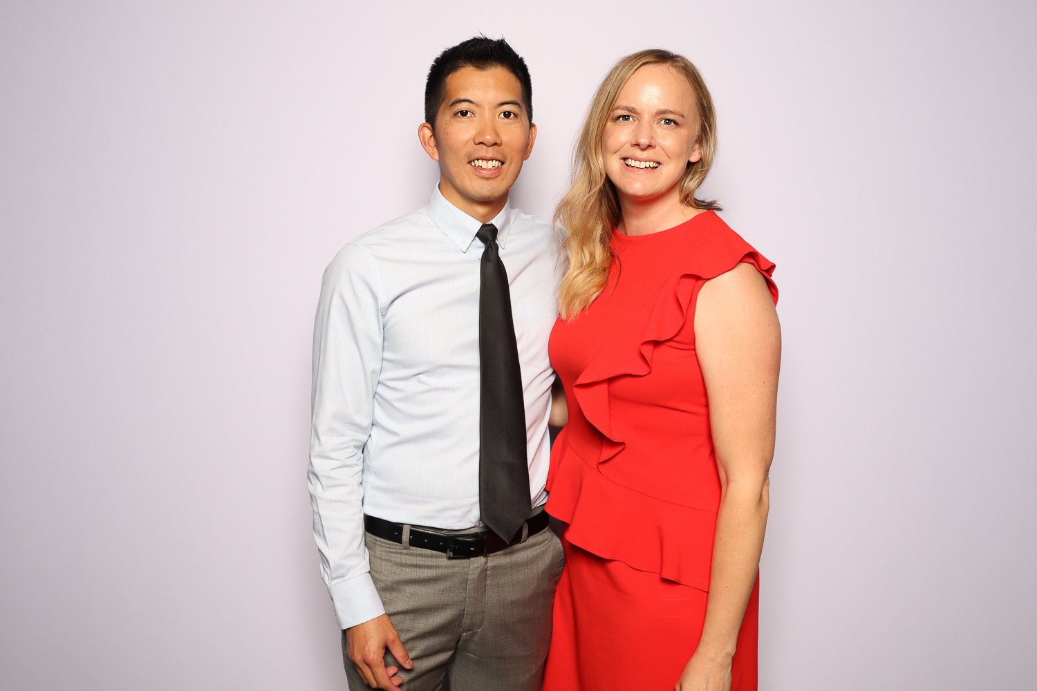 A man and woman posing together, both smiling, with the man dressed in a white shirt and black tie, and the woman wearing a red dress with ruffle details.