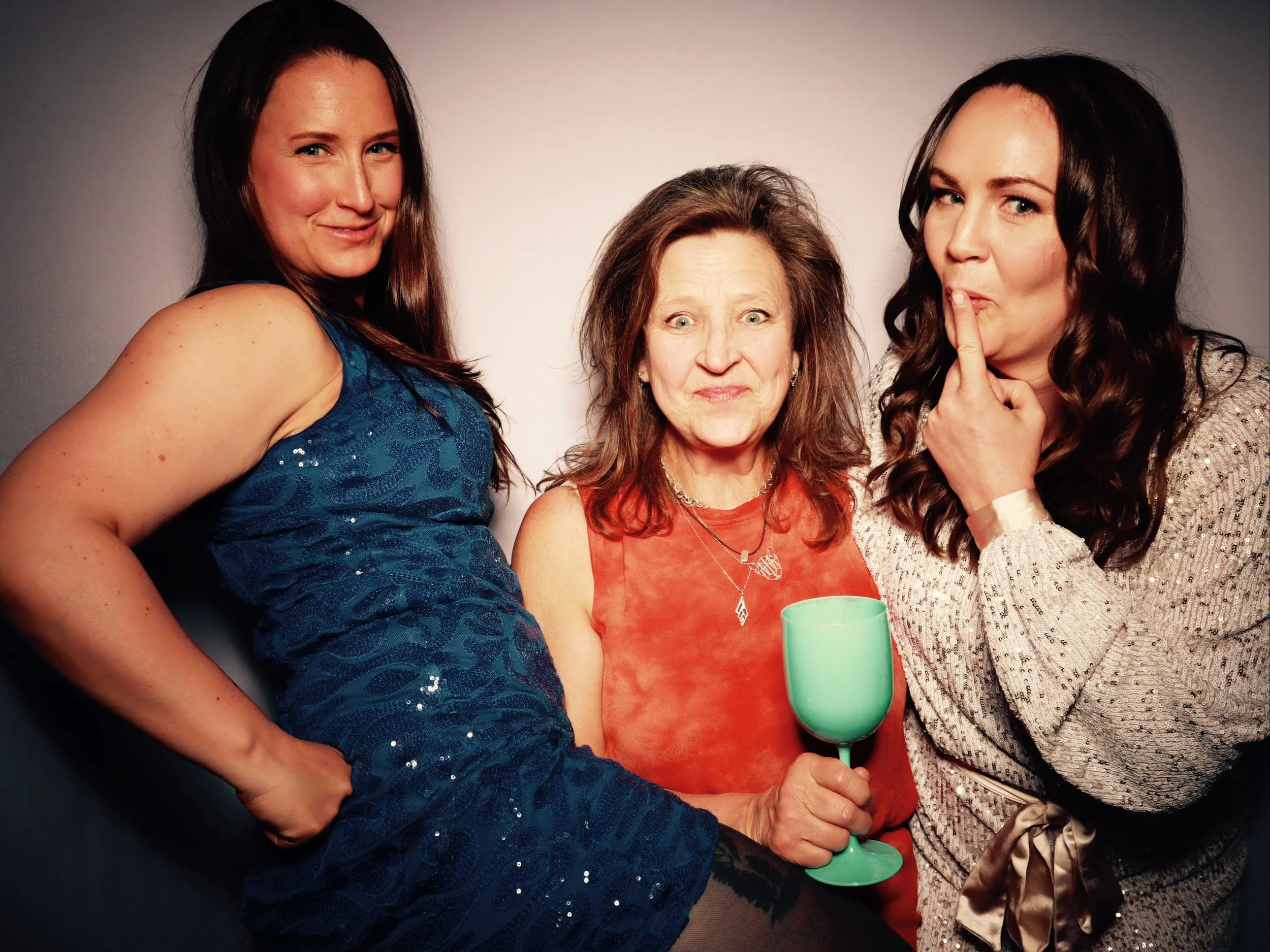 Three women posing together at a party. The woman on the left is wearing a blue dress, the woman in the middle is holding a green wine glass and making a surprised face, and the woman on the right is wearing a sparkly silver top, shushing with her fi