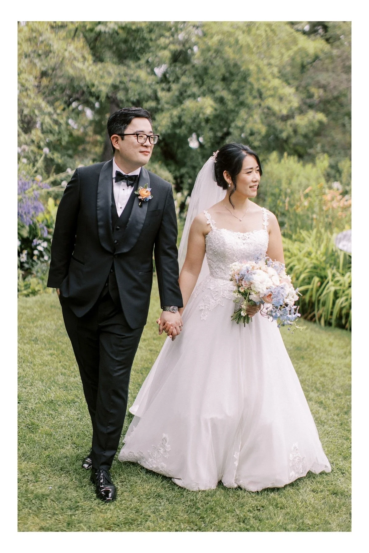 A bride in a long wedding dress holds her bouquet, hand-in-hand with the groom wearing a black tuxedo, as walk through the gardens of the Calgary Zoo to their wedding ceremony