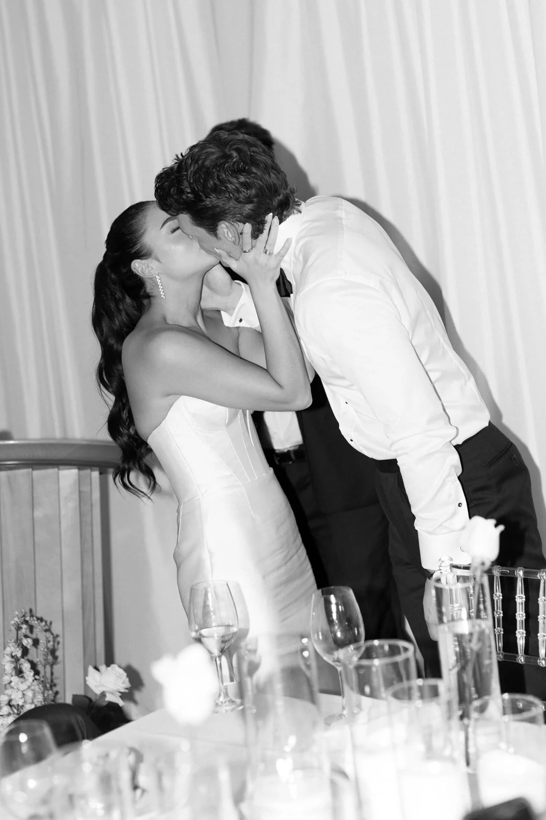 A couple kissing at a wedding reception, with the woman in a strapless wedding dress and the man in a white shirt.