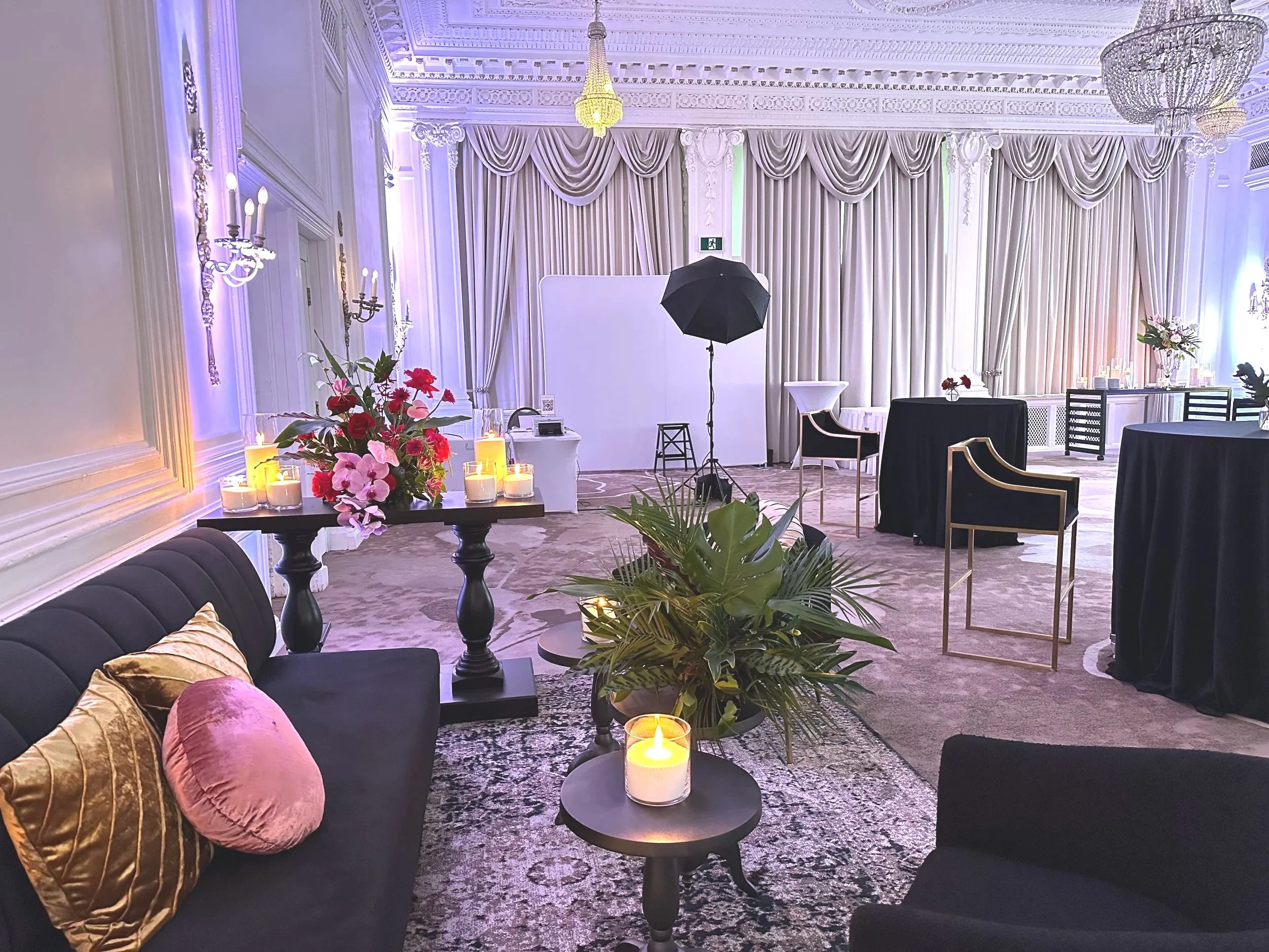 Elegant event space with high ceilings, ornate moldings, and large curtains. Decor includes floral arrangements, candles, seating with velvet pillows, and photography equipment like a studio light. The room is softly lit with chandeliers and wall sconces.