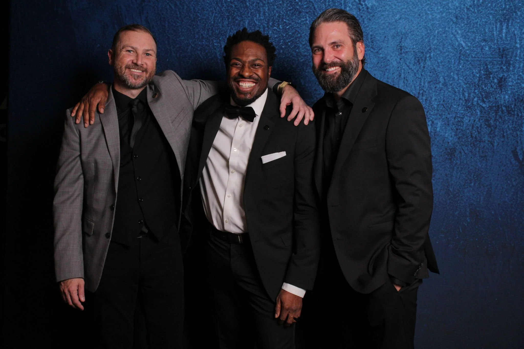 Three men in formal attire standing against a blue textured background, smiling, with arms around each other's shoulders.