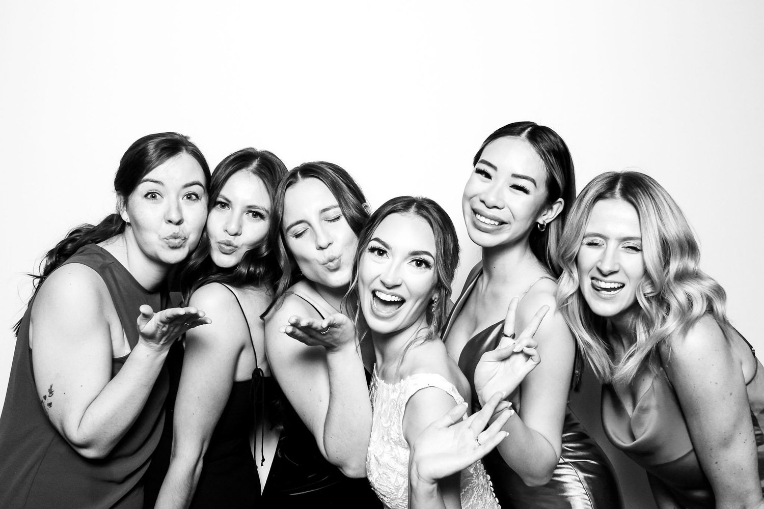 A group of six women in stylish dresses posing and making playful gestures, smiling and blowing kisses in a black-and-white photo.