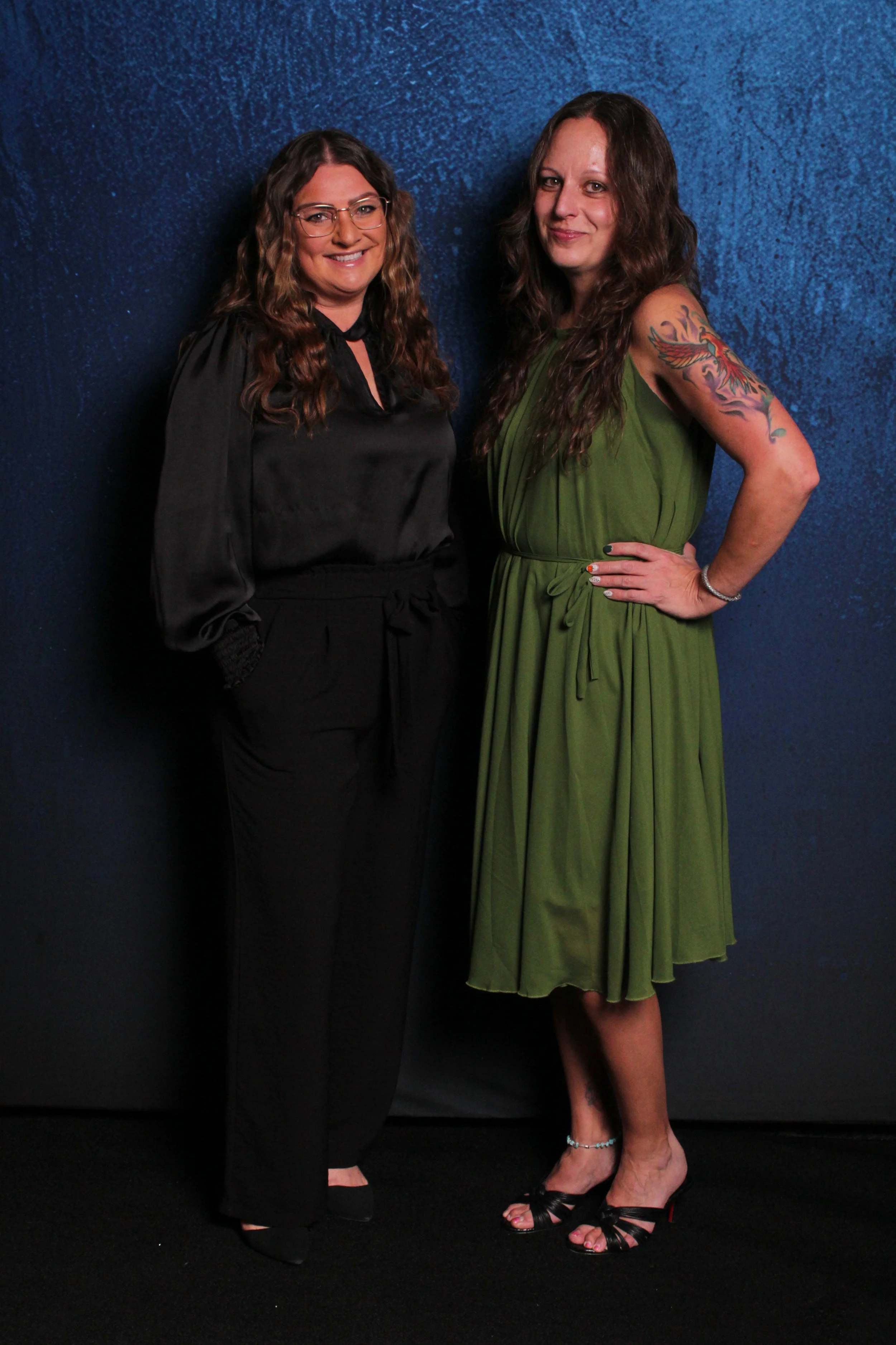 Two women standing side by side against a blue textured background, smiling at the camera. The woman on the left is wearing a black outfit with glasses and has long wavy brown hair. The woman on the right is wearing a green dress with a tattoo on her