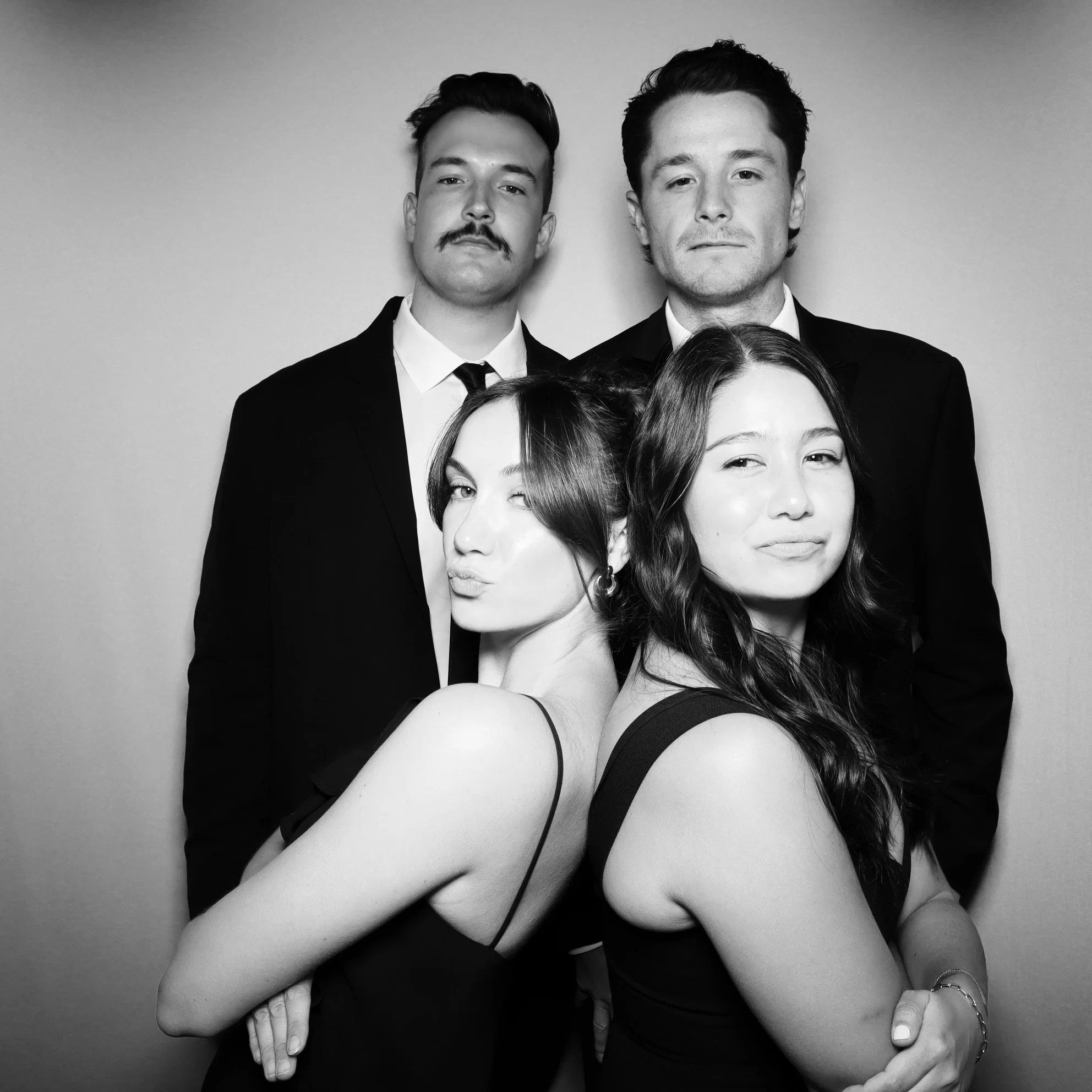 Black and white photo of four young adults, two men in suits in the back, two women in sleeveless dresses in the front, posing confidently.