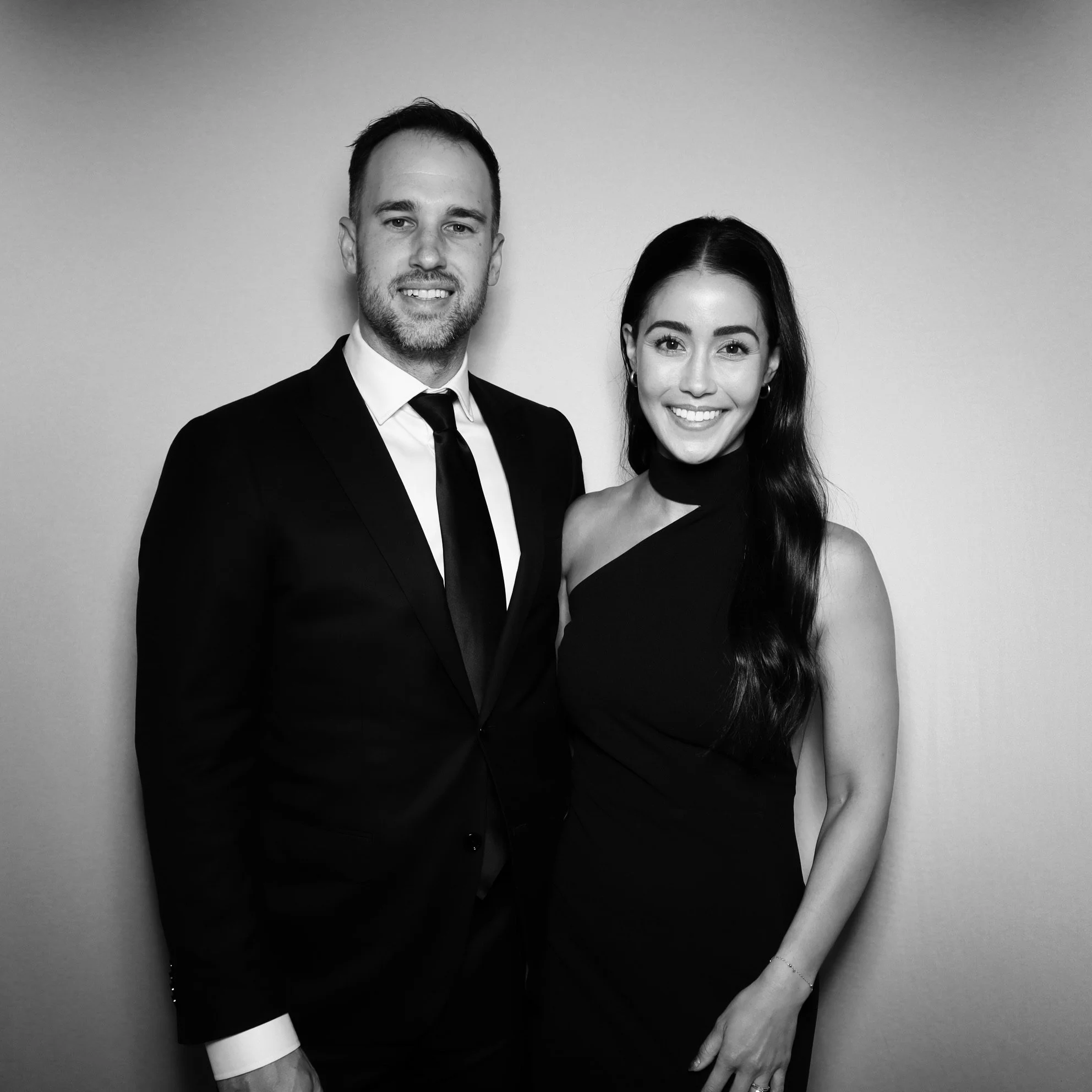A man and woman dressed in formal attire, smiling and standing close together against a plain background.