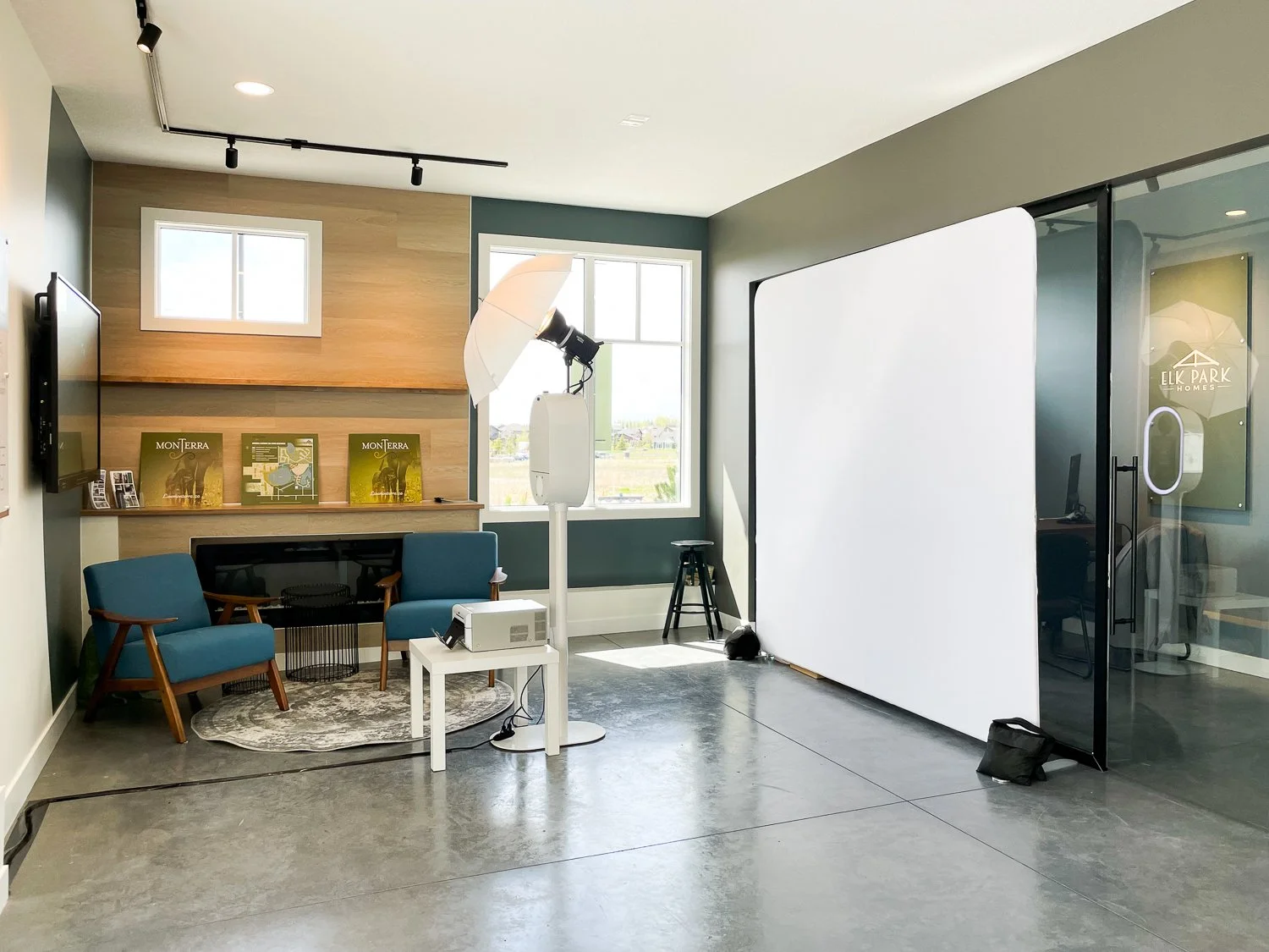 A professional photography studio setup with a white backdrop, lighting equipment including an umbrella light, seating area with two blue chairs, a small round table, and a television mounted on the wall.