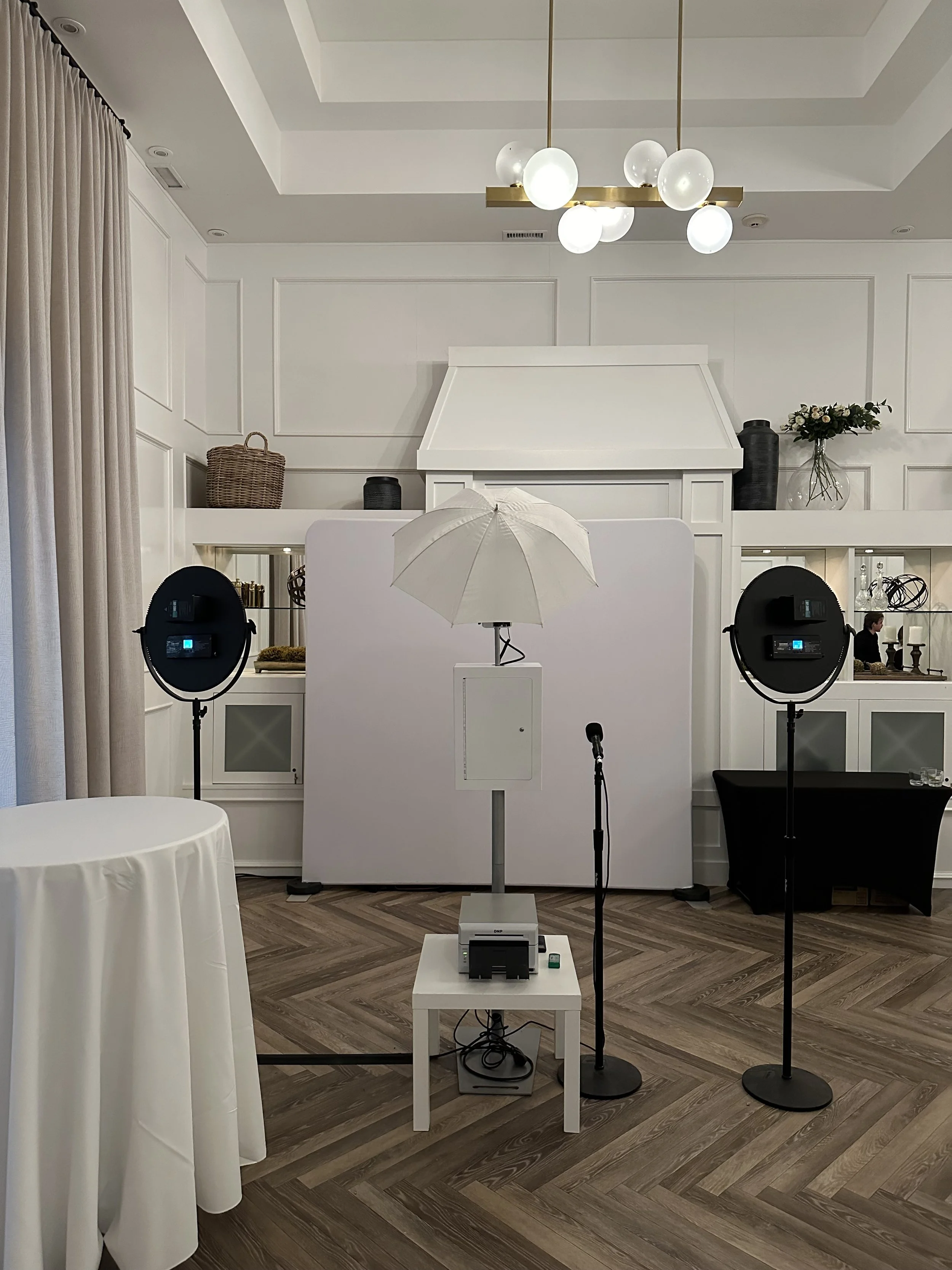 Photo of a professional photography or videography setup with studio lights, a backdrop, and a camera on a tripod inside a room with white walls and wooden flooring.