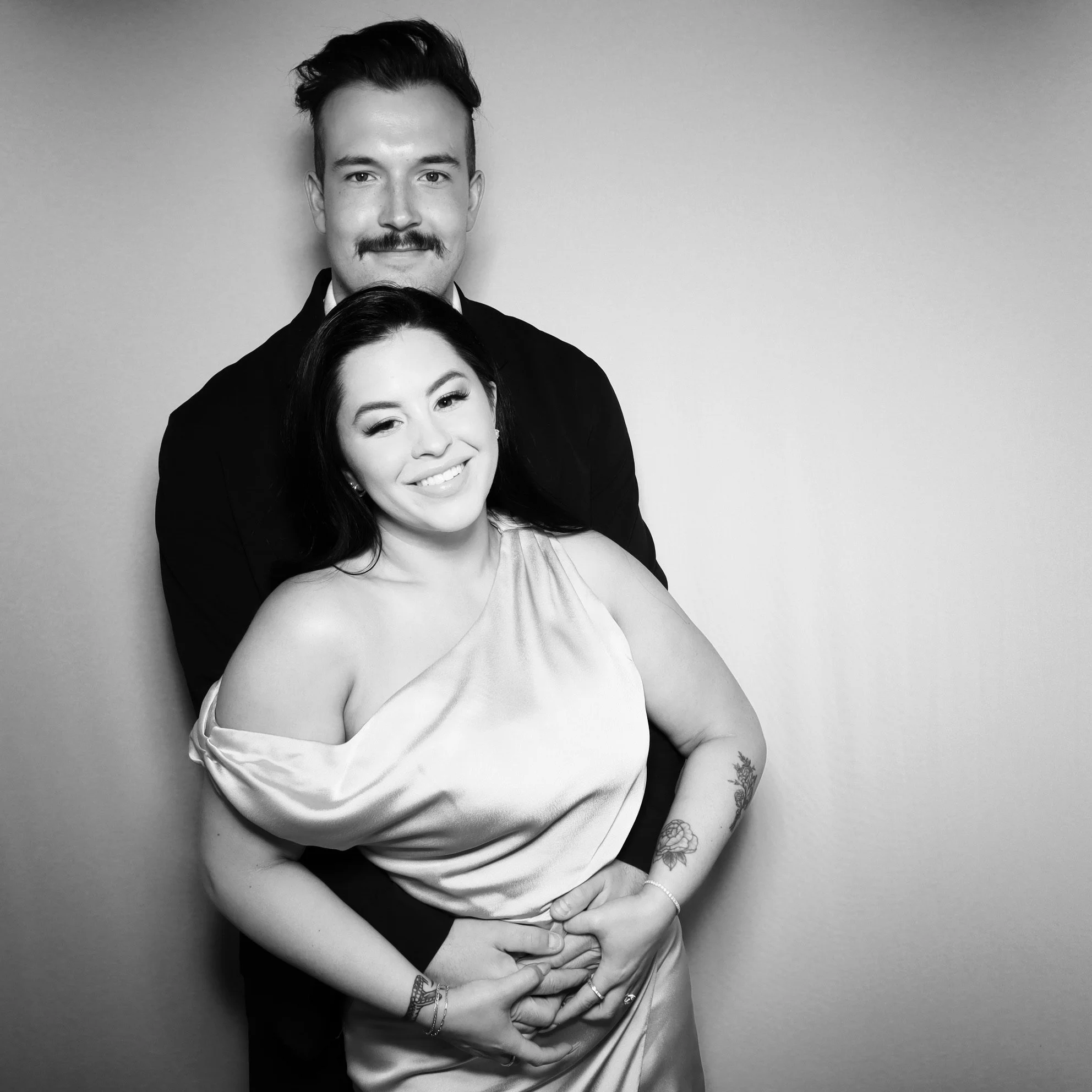 A happy couple in formal attire posing against a plain background, with the man standing behind the woman, both smiling, with the woman winking.