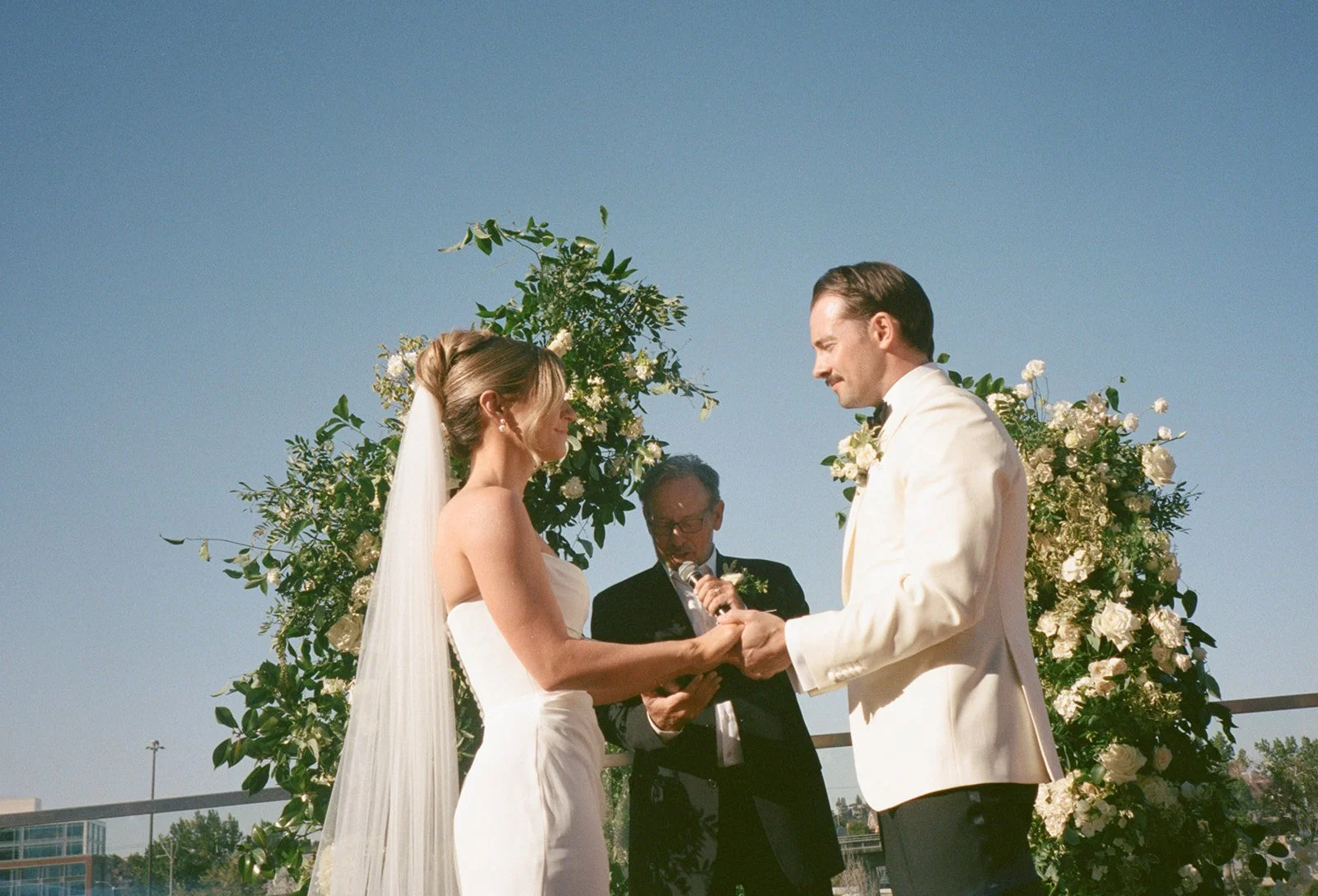 A bride and groom holding hands during their outdoor wedding ceremony under a floral arch with a clear blue sky.