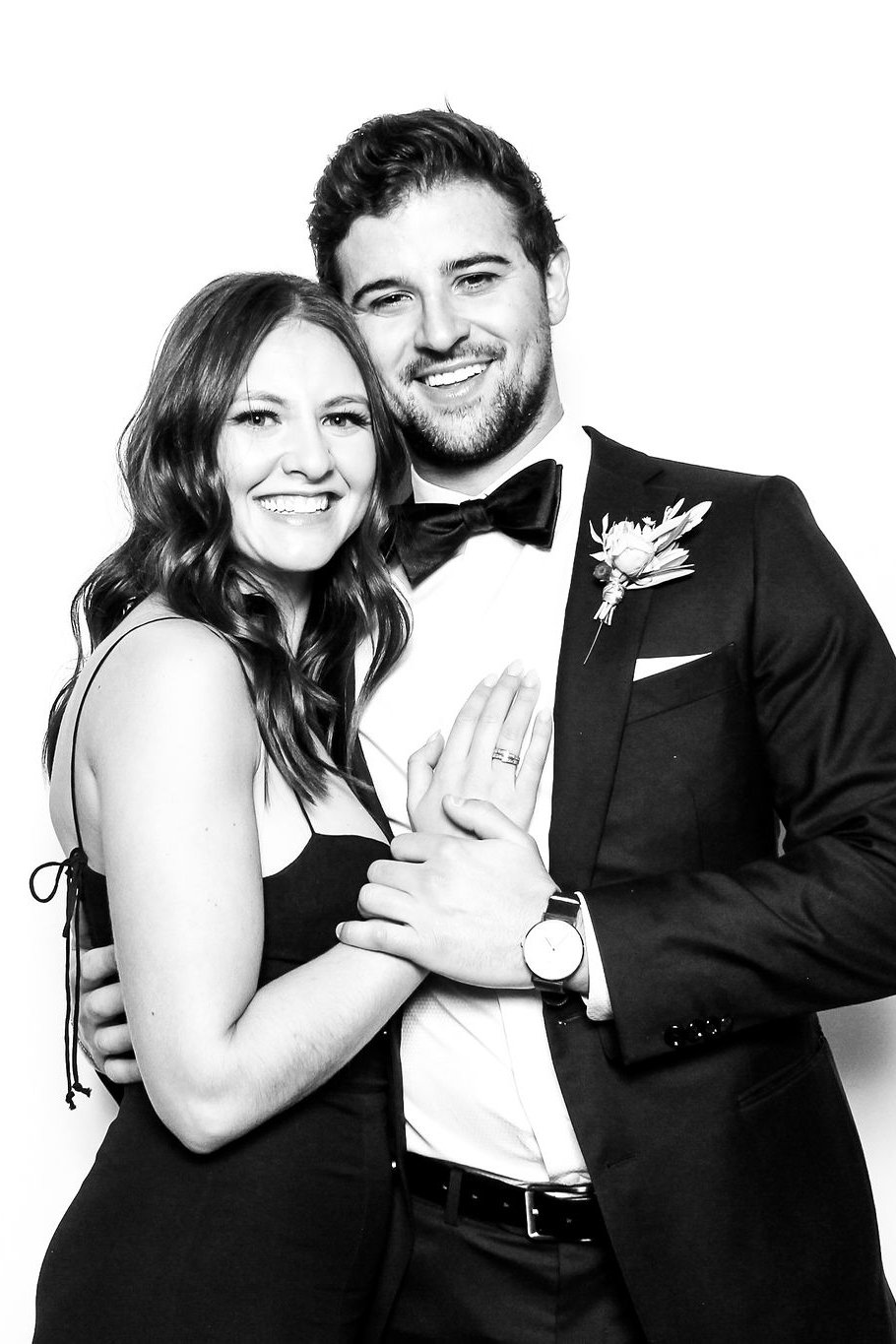 A black and white photo of a happy couple dressed in formal attire, smiling and holding hands, with the woman's hand showing a wedding ring.