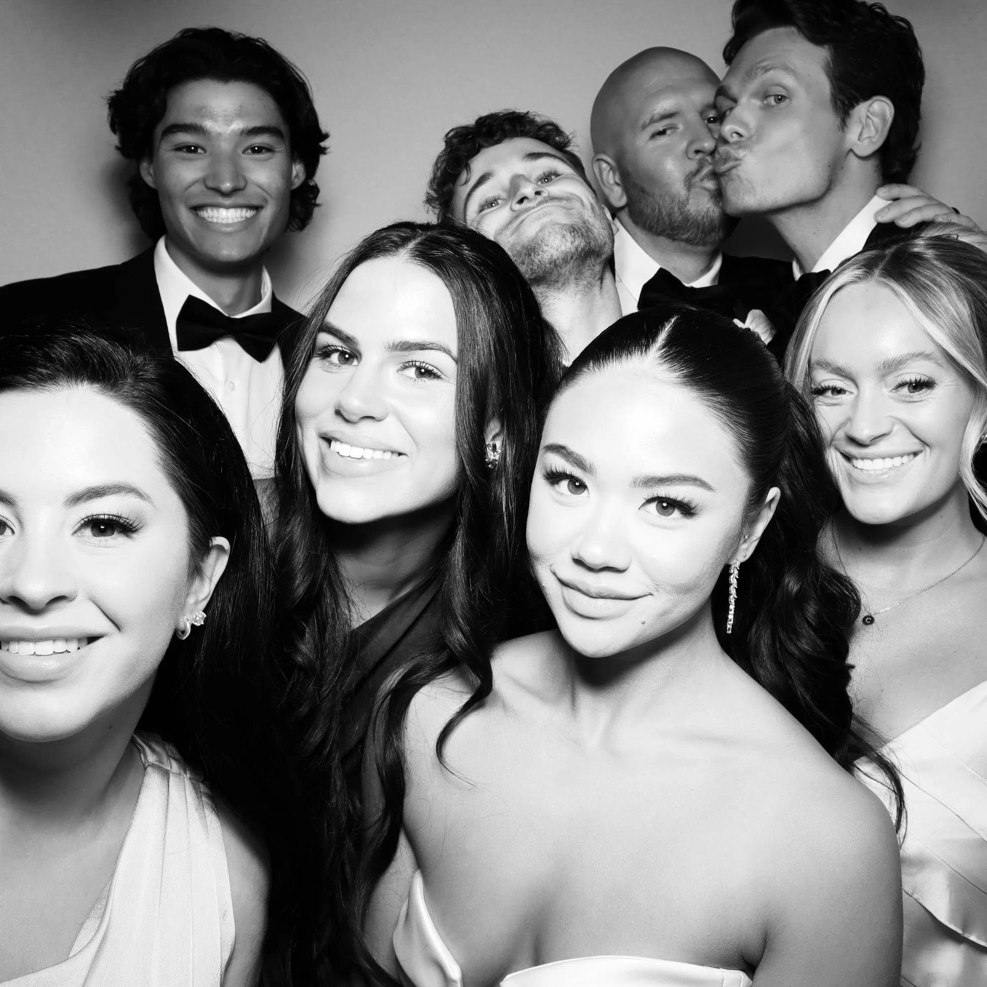 Group of six men and women dressed in formal attire, posing and kissing in a photo booth, black and white.