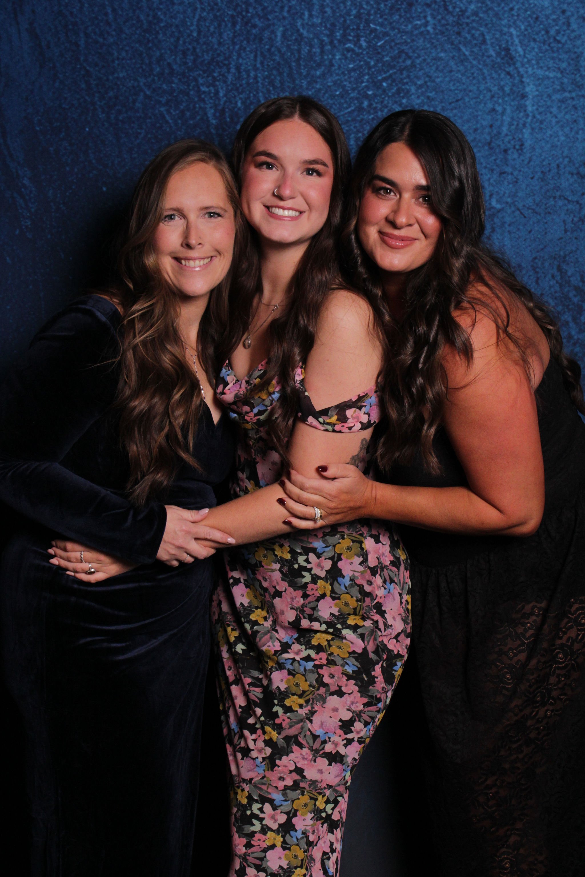 Three women are smiling and posing together in front of a blue backdrop. They are wearing elegant dresses and have long, wavy hair.
