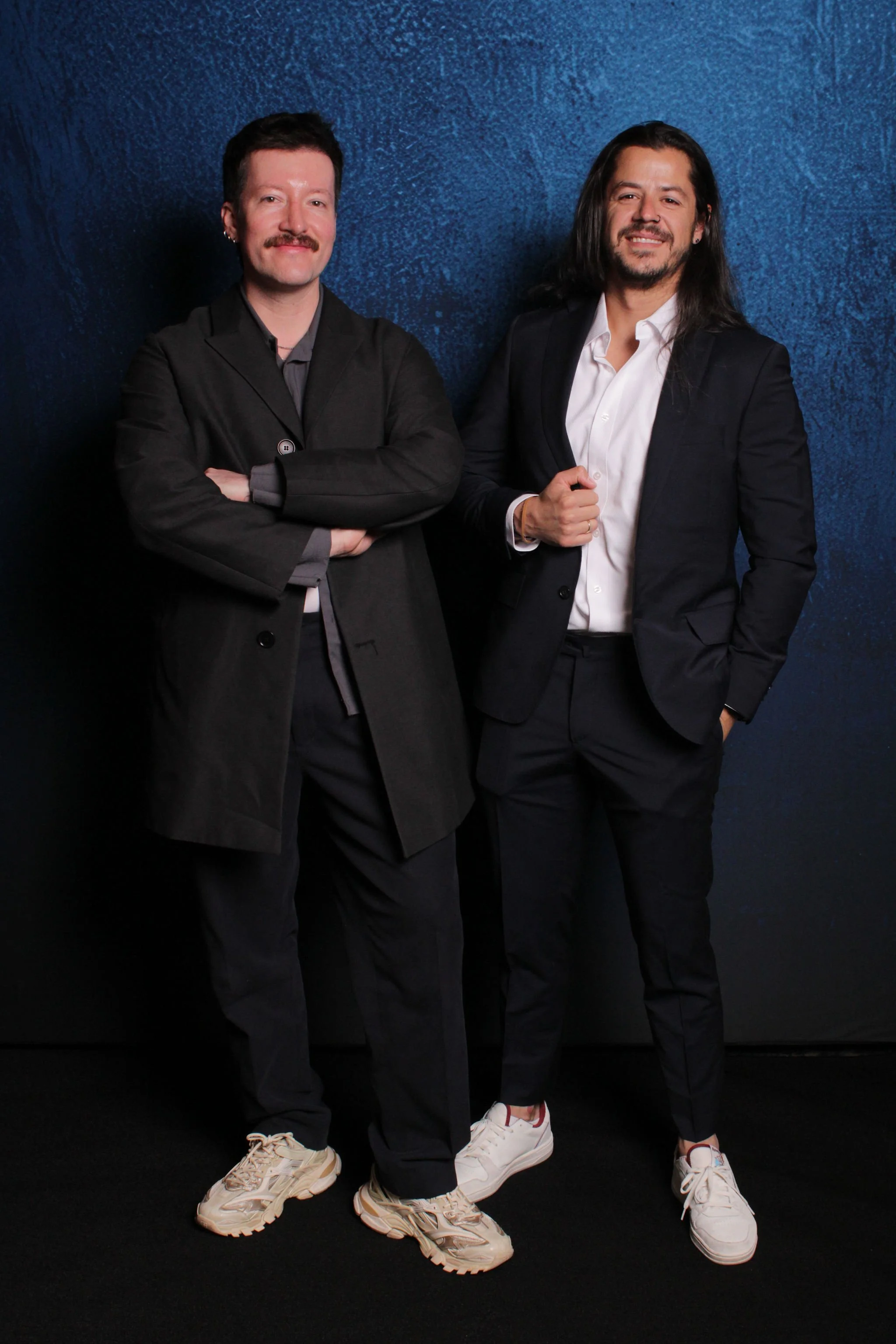 Two men standing against a dark blue textured background, one with arms crossed and the other with one hand in pocket and the other adjusting his sleeve. They are dressed in black suits and casual shoes, smiling at the camera.