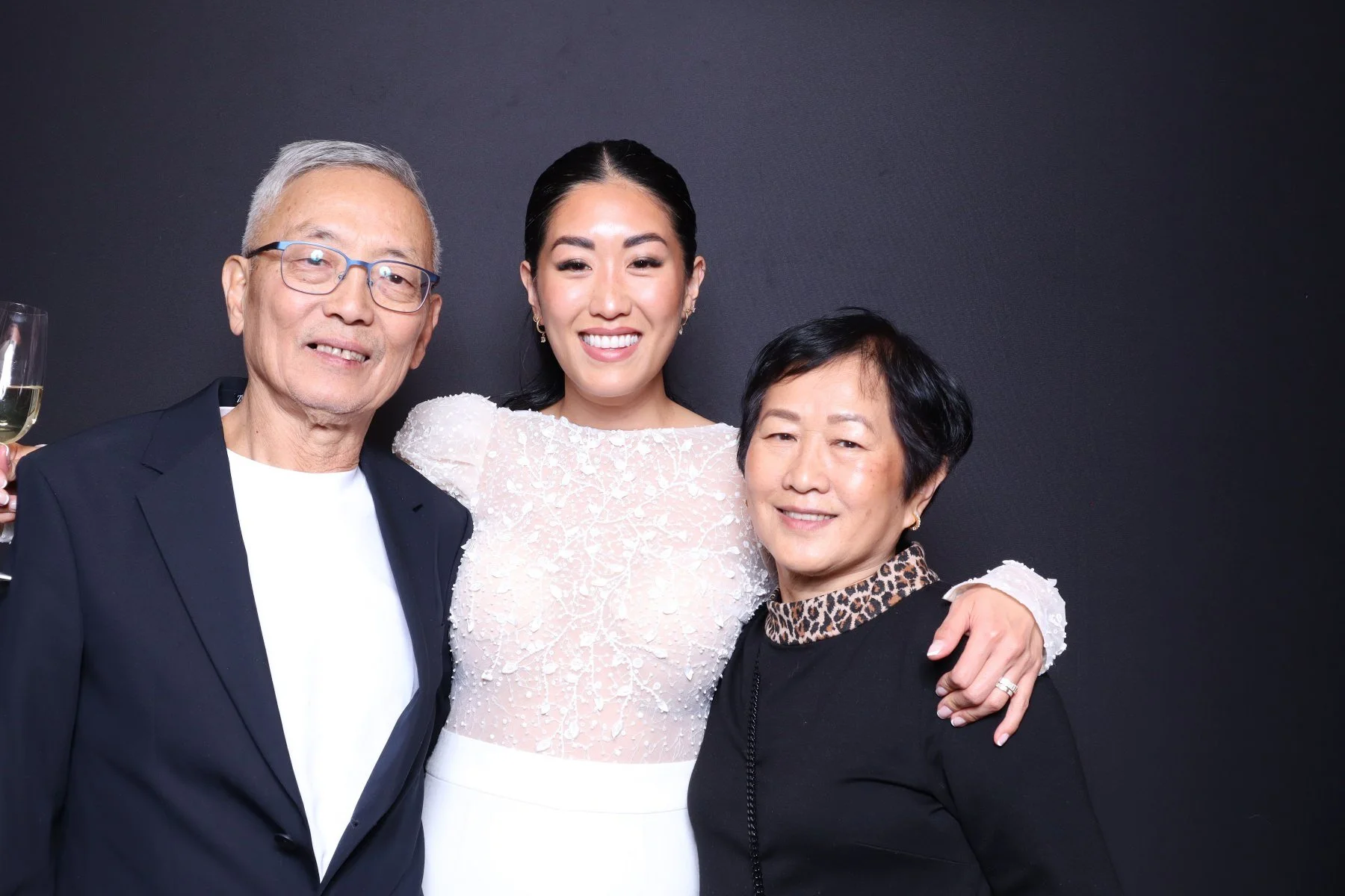 Three people, an elderly man, a woman with dark hair, and an older woman, are smiling and posing together against a black background. The elderly man is wearing glasses, a black suit jacket, and a white shirt, and is holding a glass of champagne. The