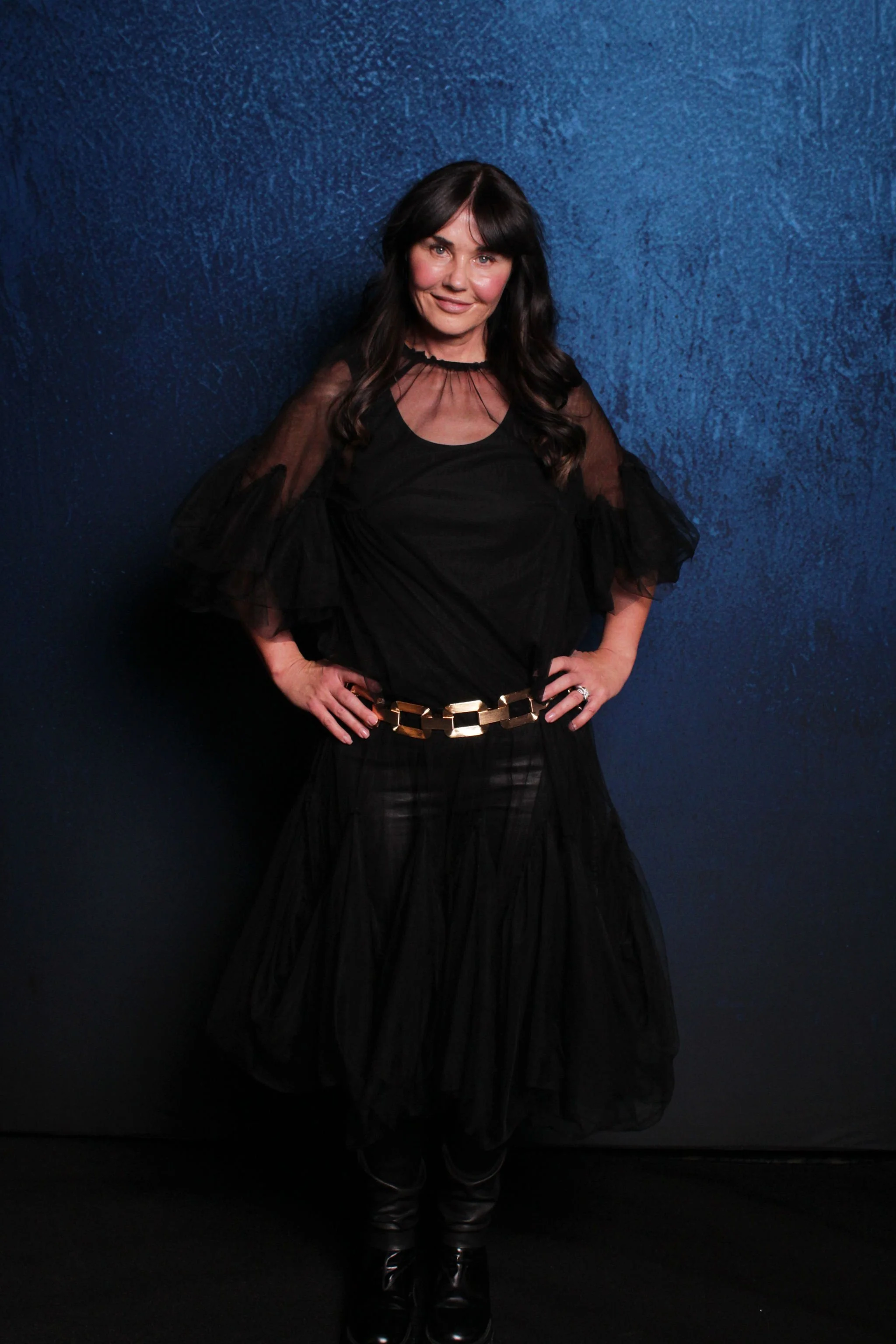 A woman standing in front of a textured blue wall, wearing a black blouse with sheer sleeves, a black skirt, black boots, and a gold belt.