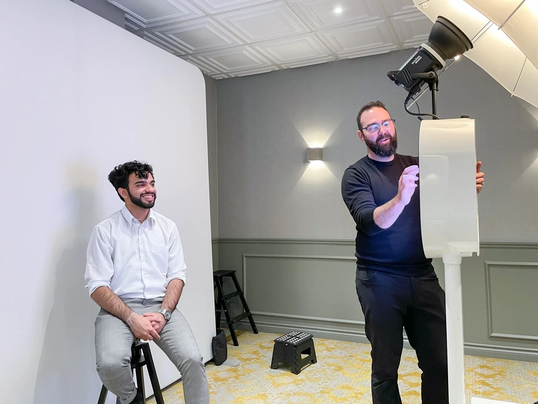 Two men in a photography studio; one sitting on a stool smiling, the other standing adjusting a white reflector, with a white backdrop and gray walls.