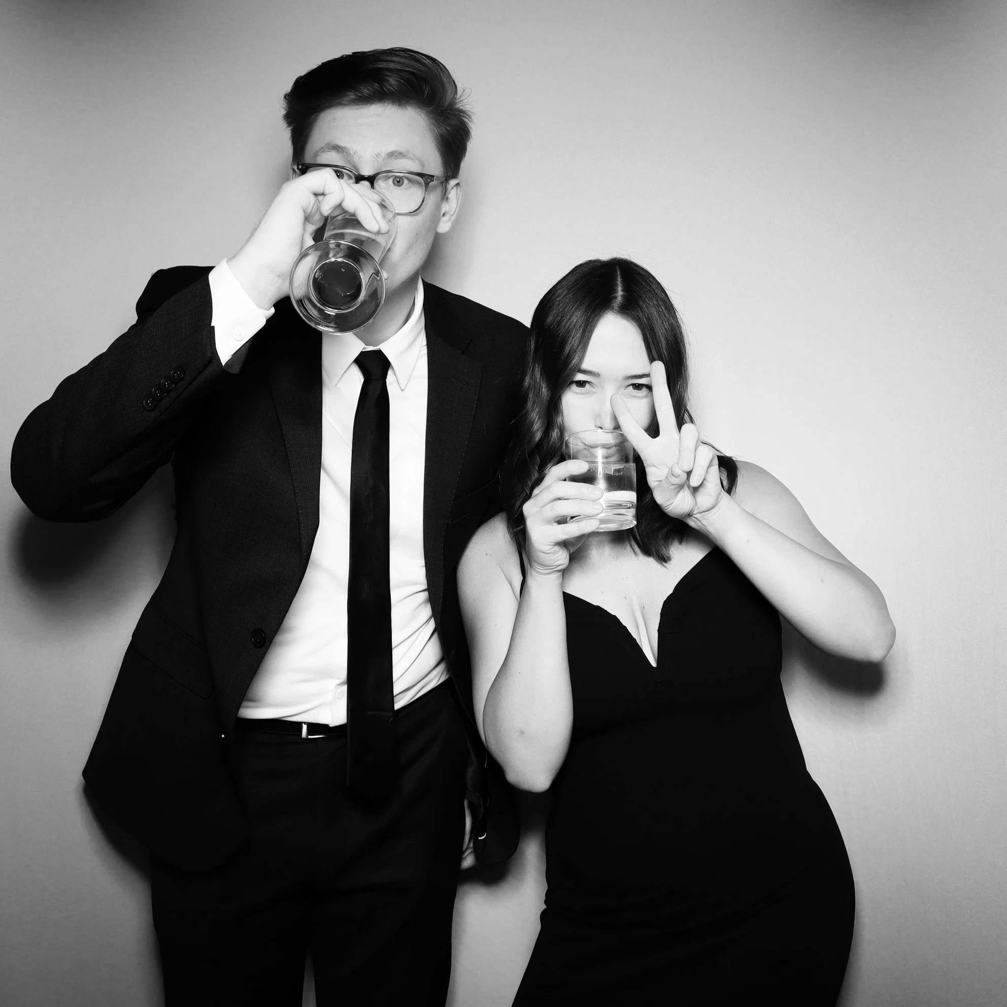 Two people, a man in a suit and a woman in a black dress, drinking and making peace signs at the camera in a photo booth.