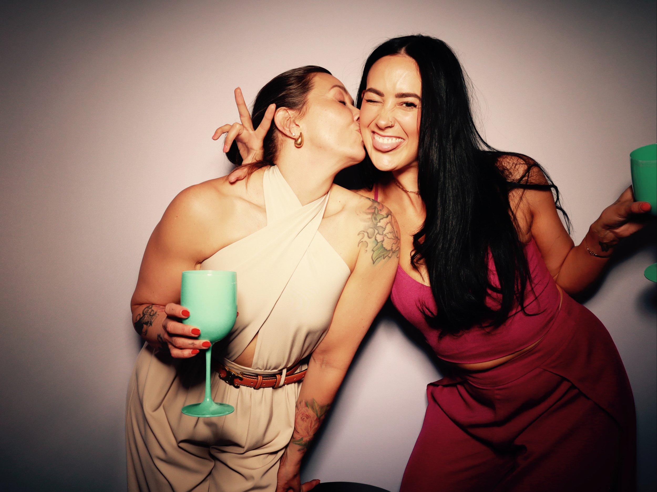 Two women celebrating, one is kissing the other on the cheek, both holding green wine glasses, smiling, against a plain background.
