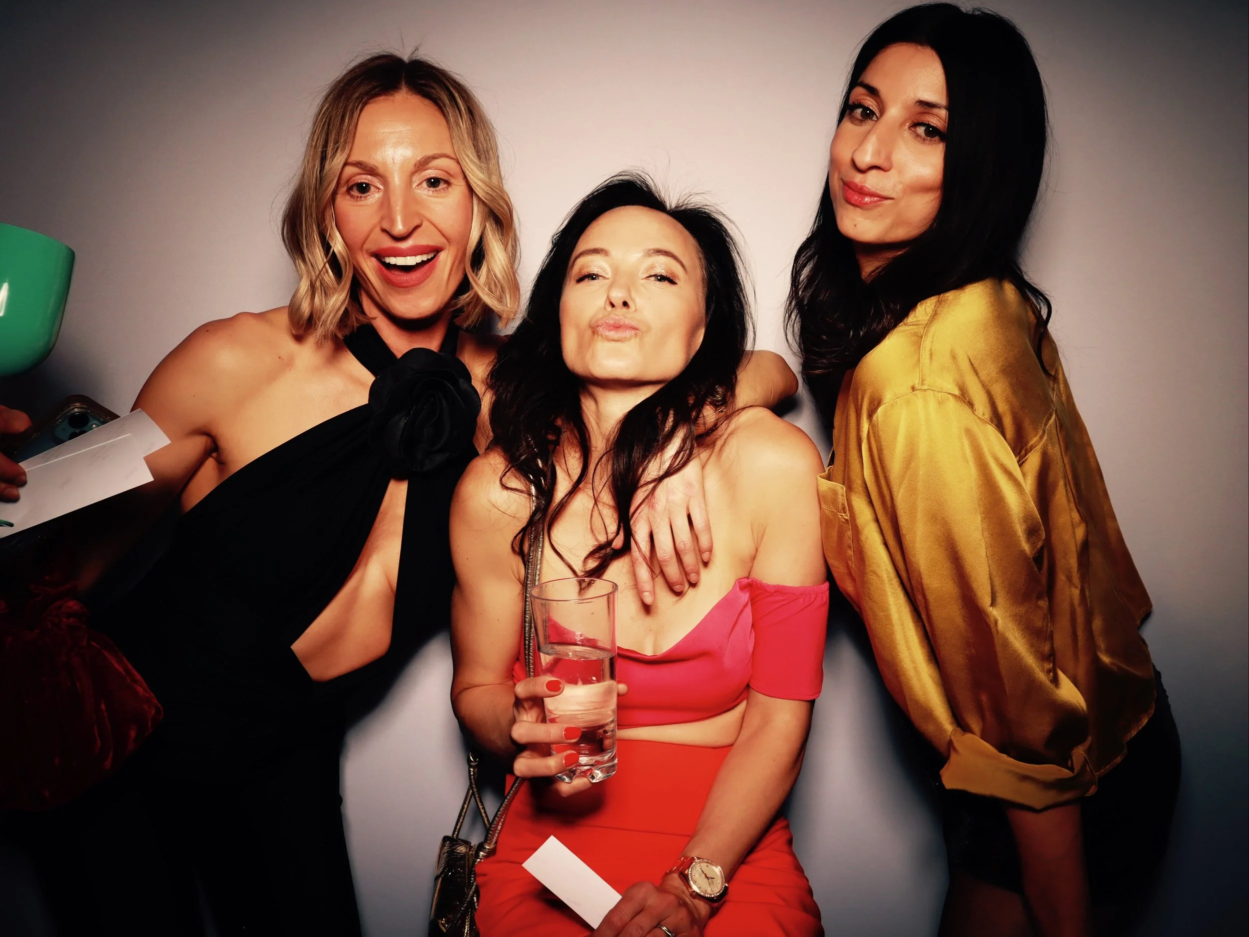 Three women at a party, smiling and posing for the camera, one holding a glass of water, with a neutral background.