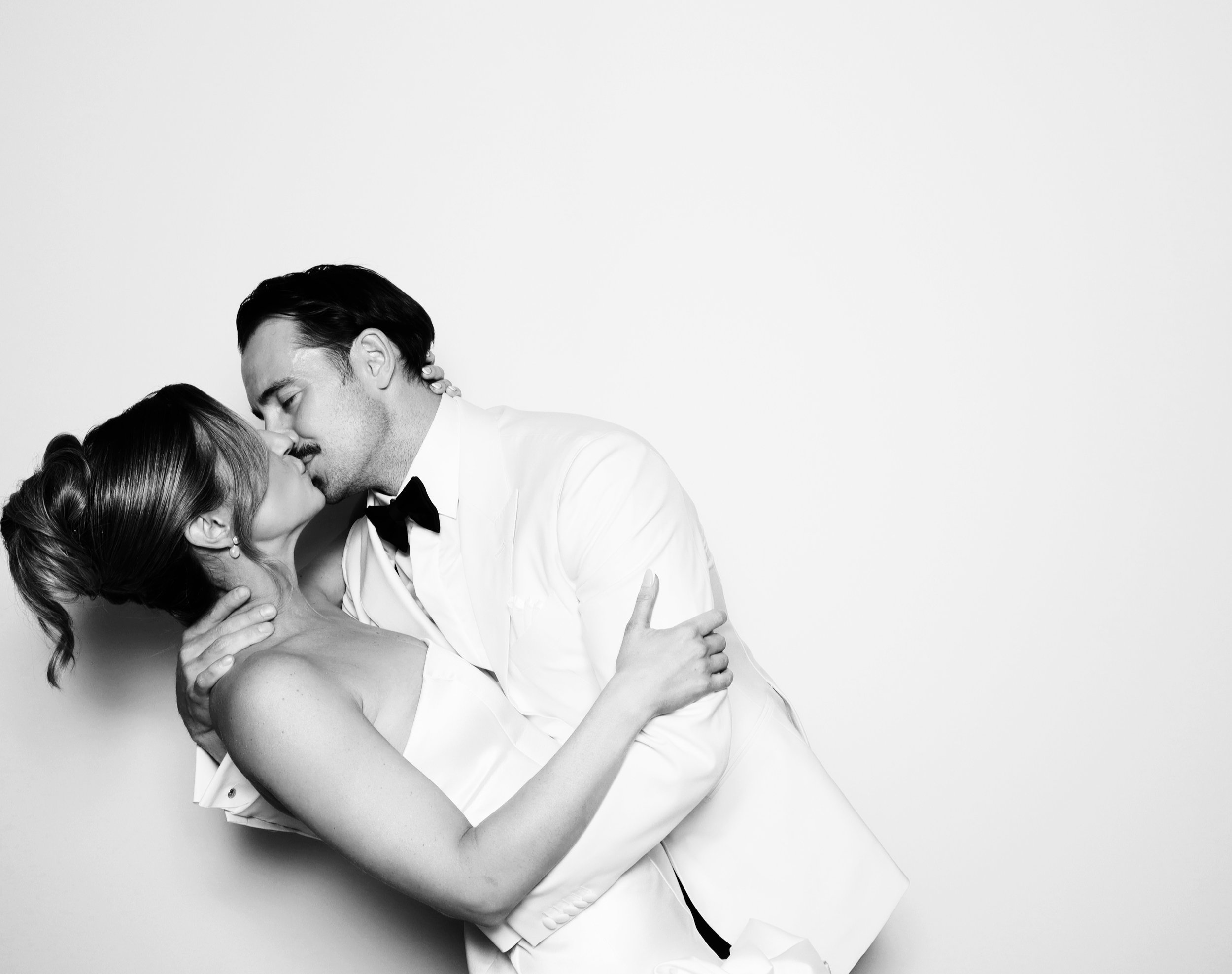 A black-and-white photo of a bride and groom kissing, with the groom dressed in a tuxedo and the bride in a wedding gown, against a plain light background.