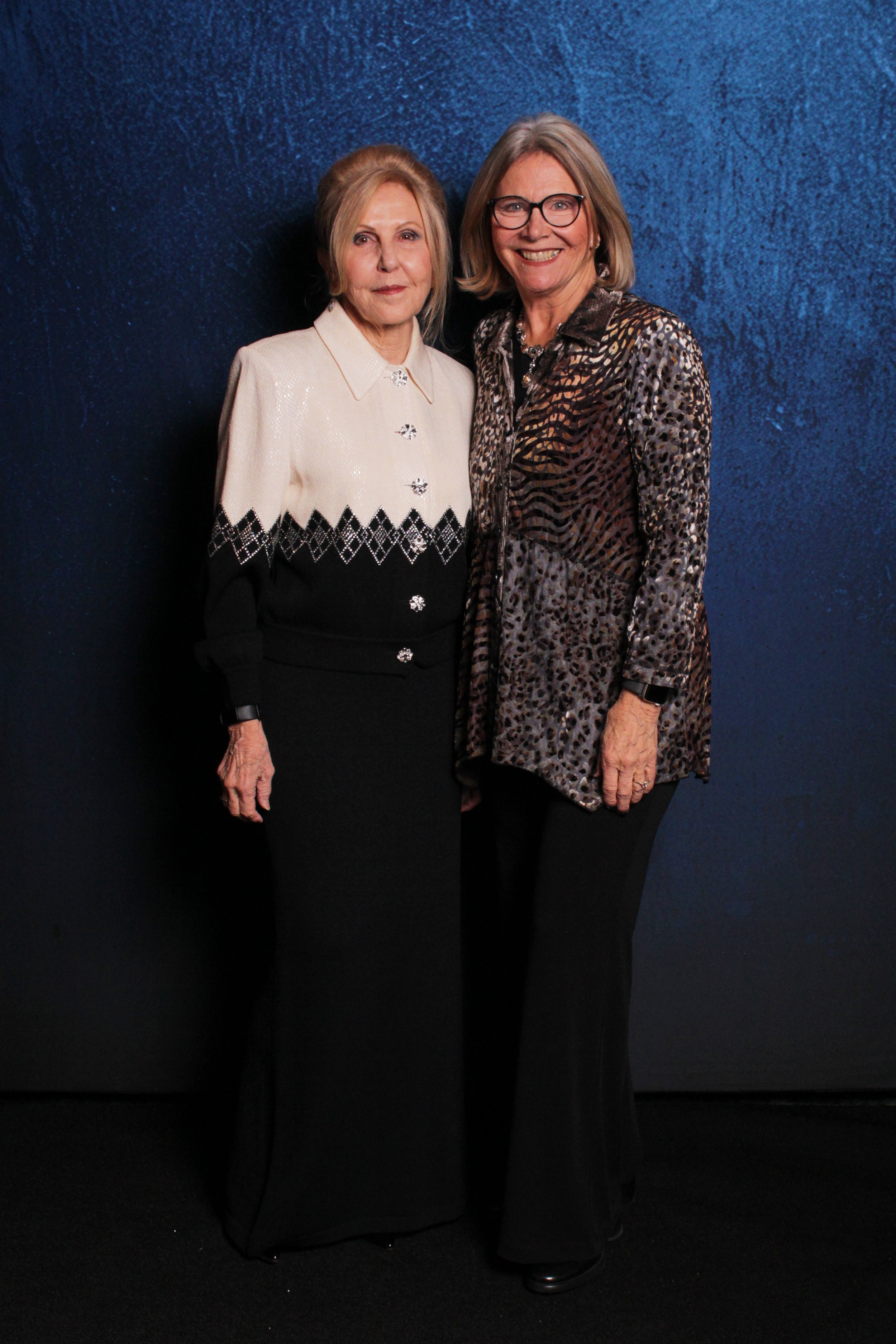 Two women standing together against a dark blue background. The woman on the left is wearing a white and black sweater with decorative buttons and a long black skirt. The woman on the right is wearing glasses, a patterned jacket with animal print, an