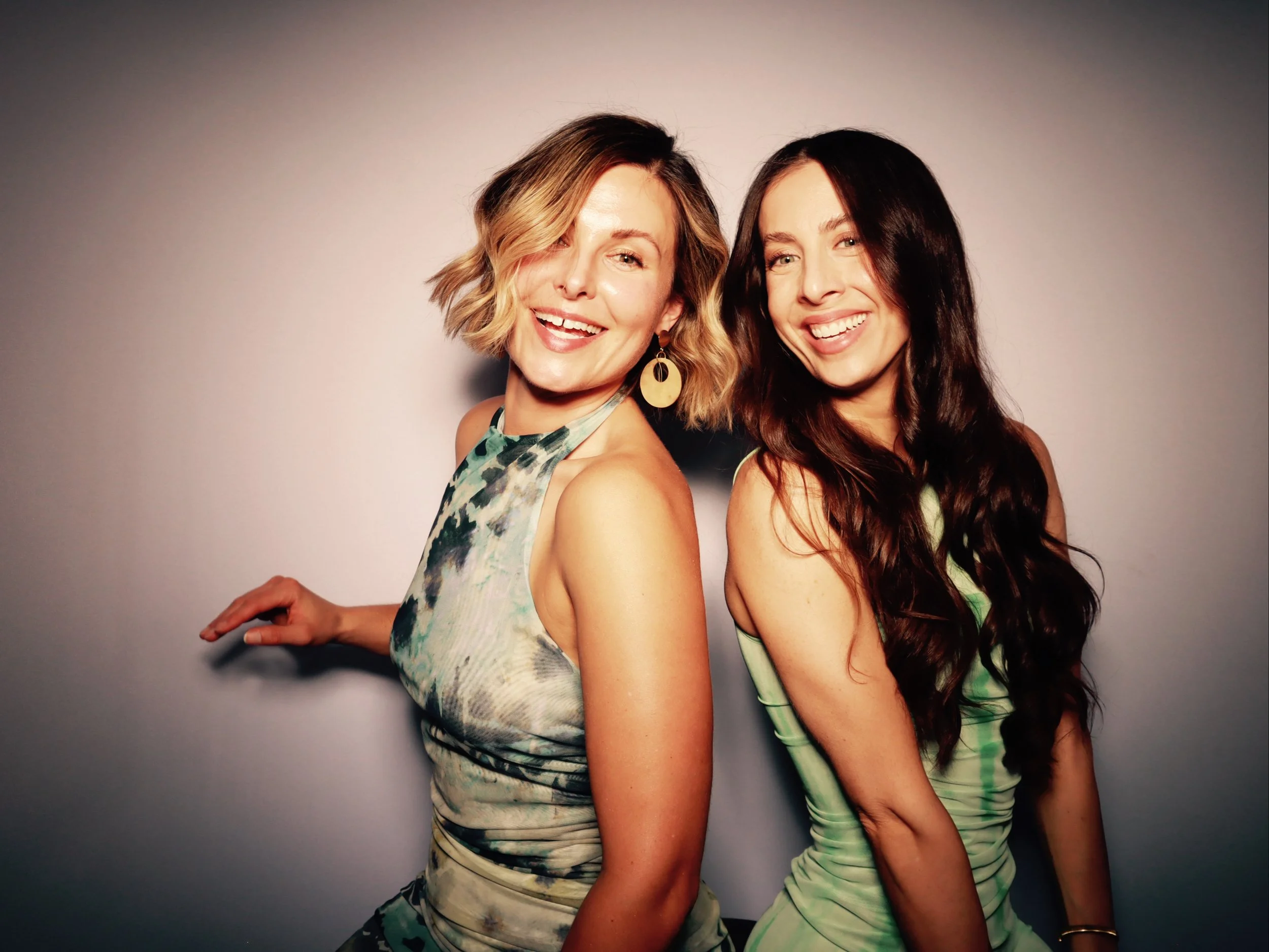 Two women smiling and posing together against a plain background, one with short wavy blonde hair wearing a sleeveless patterned dress and large earrings, the other with long wavy dark brown hair in a sleeveless green dress.