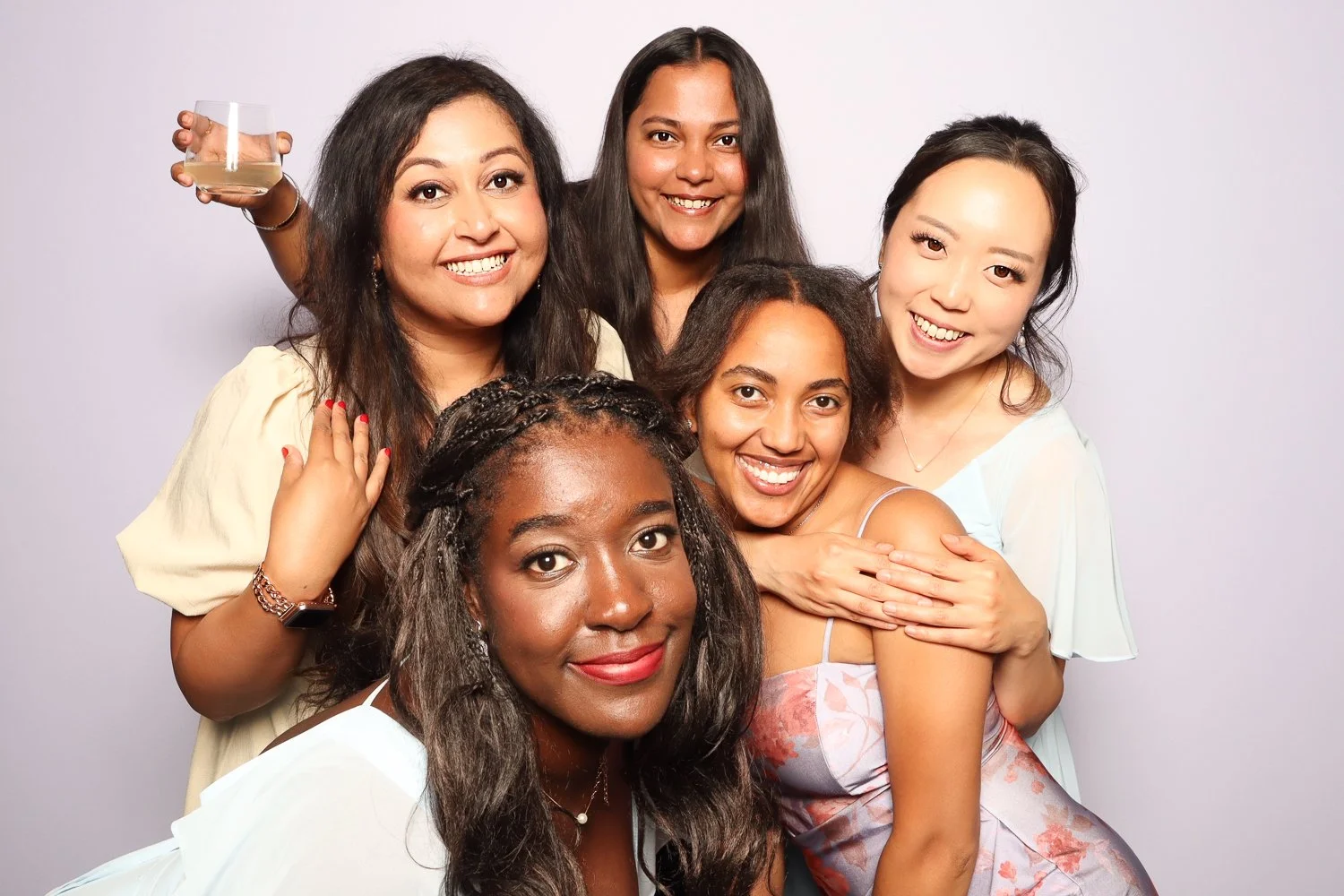Group of six women smiling and posing together against a plain background, with one woman holding a glass of white wine.