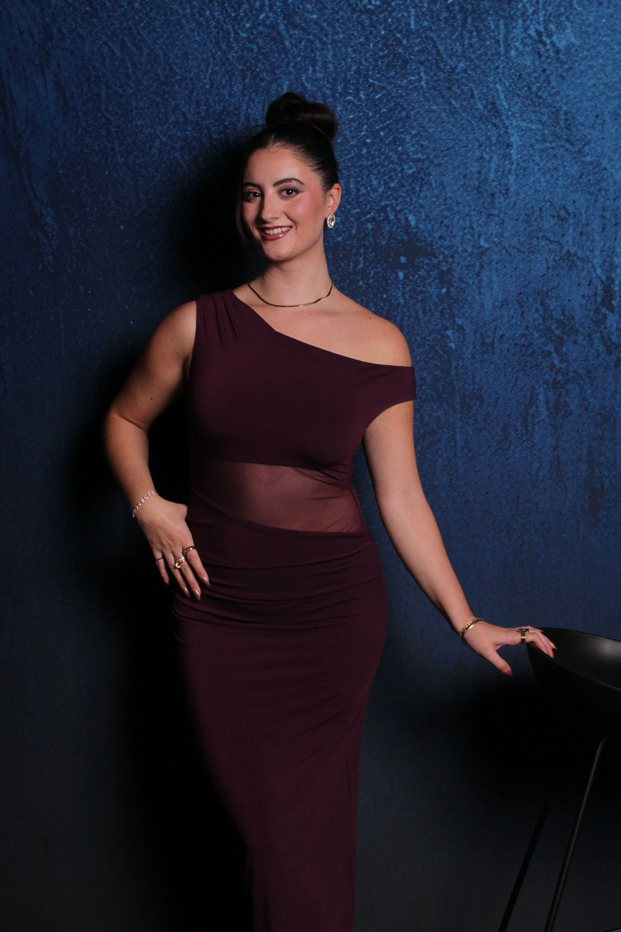 A woman in a burgundy dress with a sheer midsection, standing against a dark blue textured wall, smiling, with her hair in a bun, wearing jewelry and accessories.