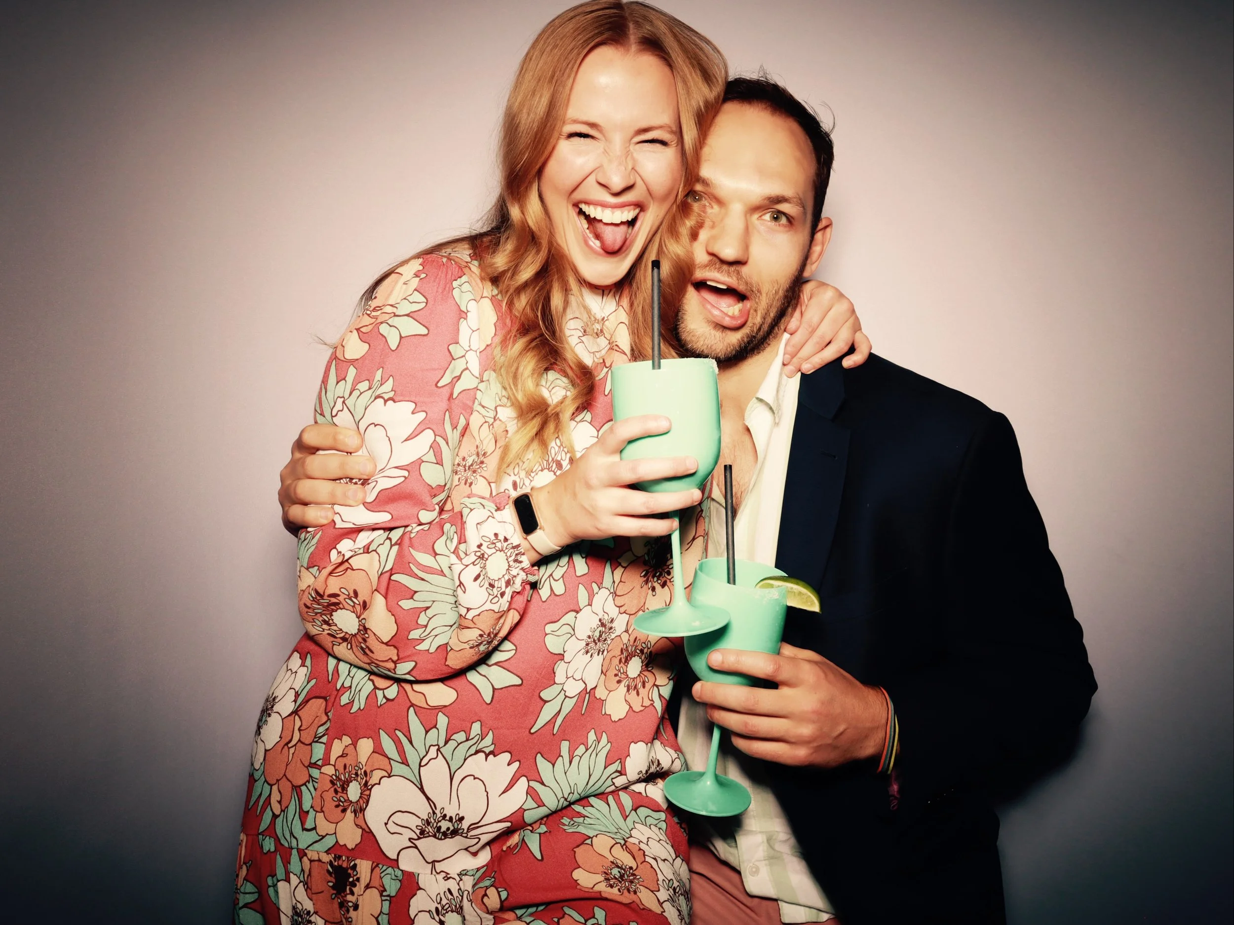 A smiling woman with long blonde hair and a floral dress embraces a man with short dark hair and a beard, both holding drinks with straws and lime wedges, against a neutral background.