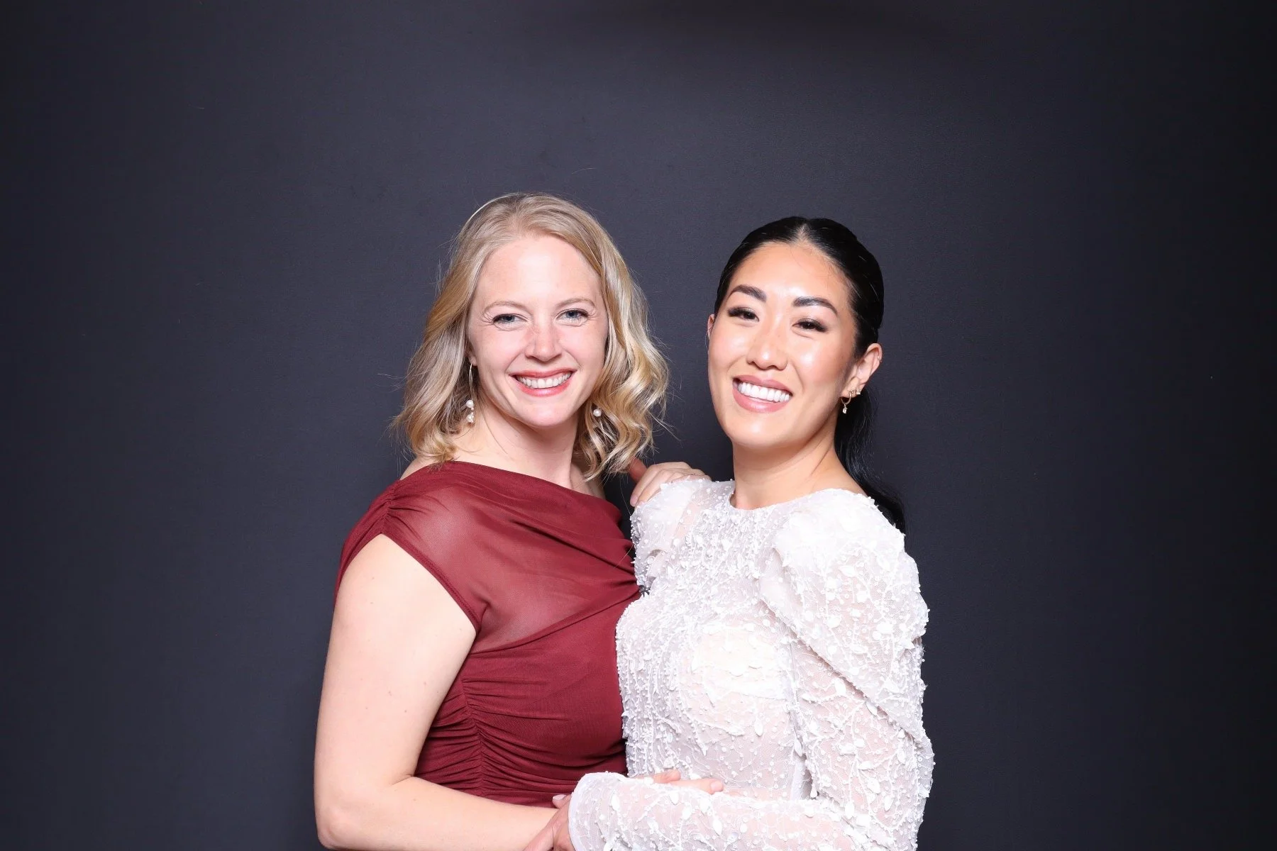 Two women smiling and posing together against a dark background, one wearing a burgundy dress and the other a white lace dress.