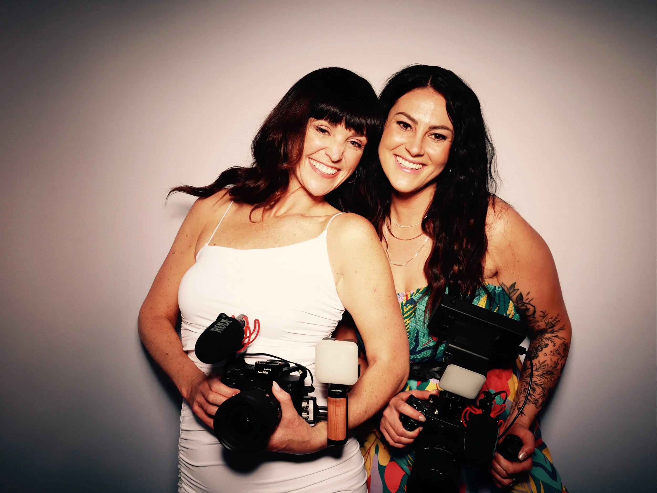 Two women smiling and posing with cameras and microphones against a neutral background.