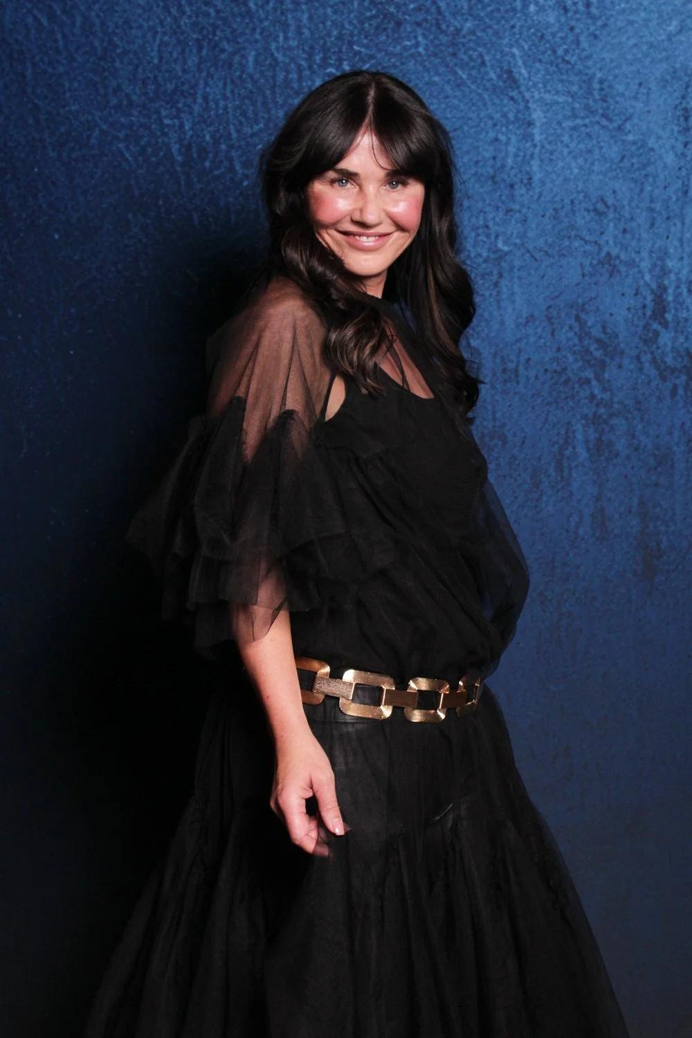 A woman with dark brown hair, smiling, wearing a black dress with ruffled sheer sleeves, a gold belt, standing against a textured blue background.
