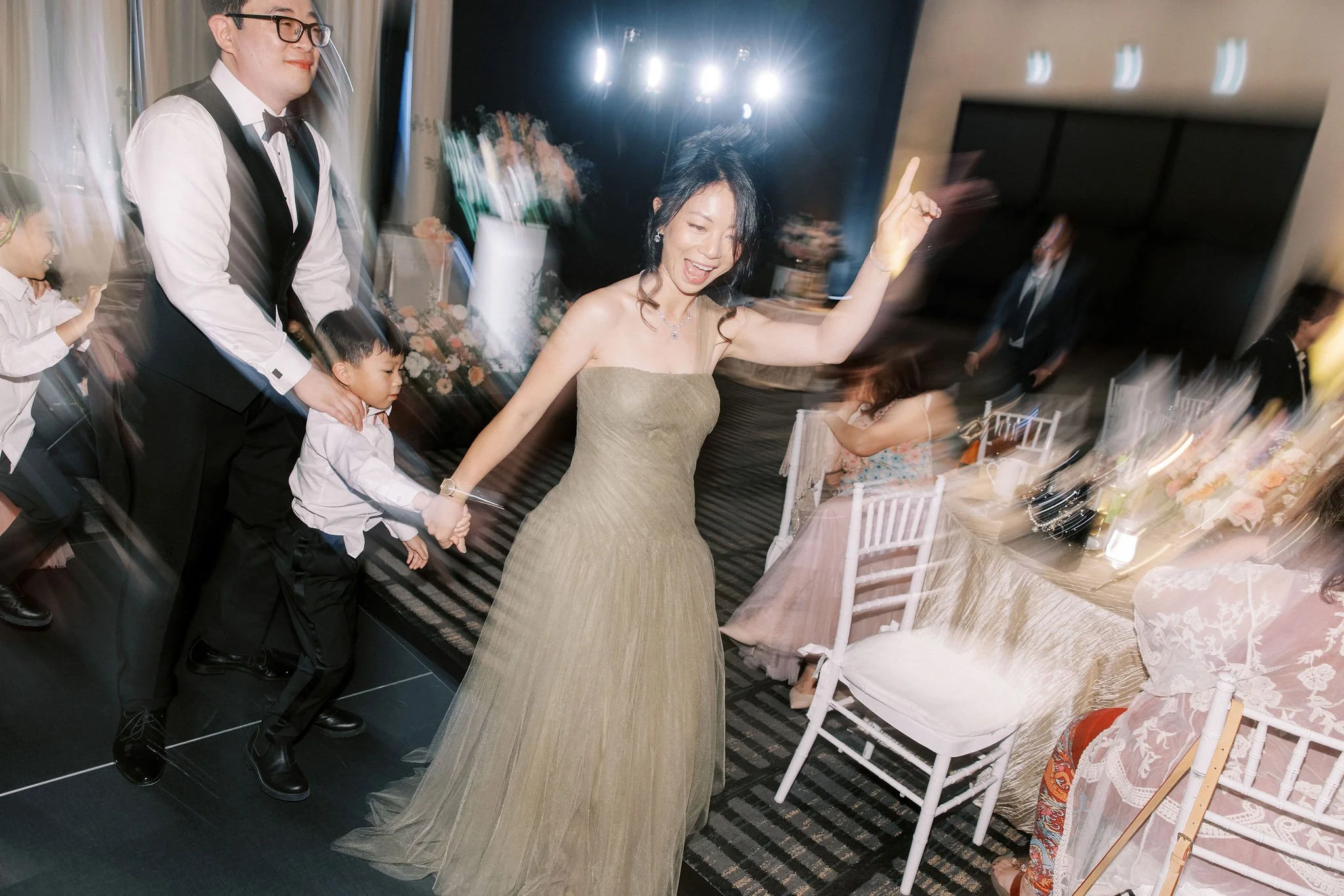 People dancing at a celebration, with a woman in a strapless gold dress, a man in a suit, and children, in a decorated venue with flowers and chairs.